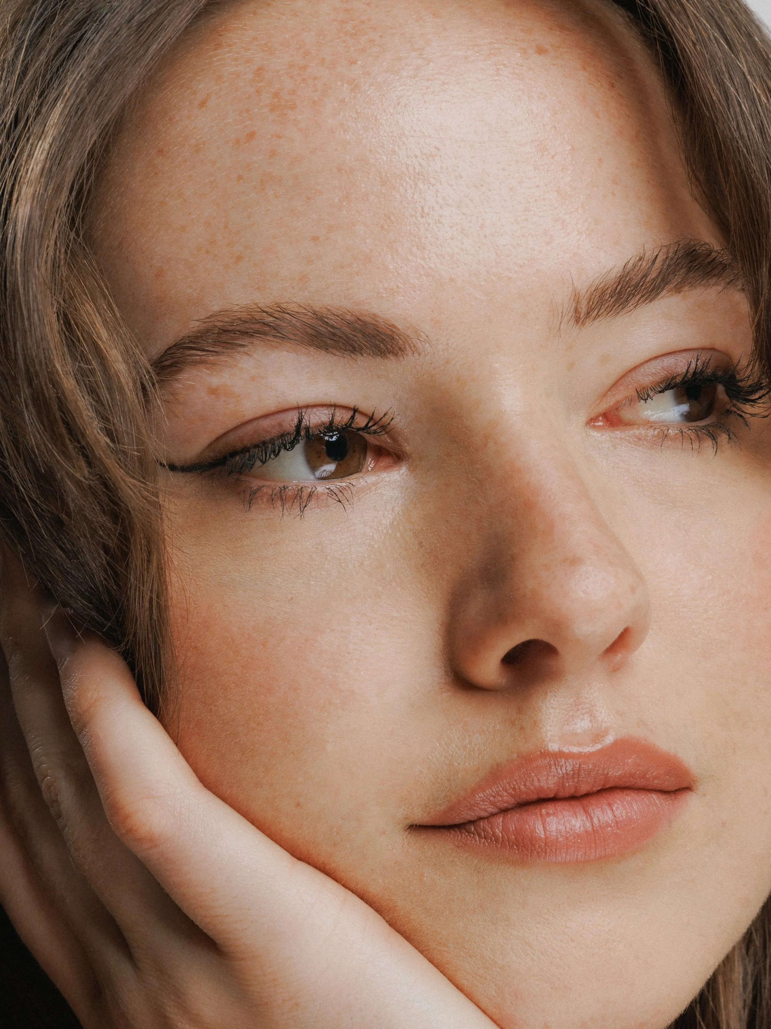 Close-up of a woman's face, showing her eye makeup, natural eyebrows, freckles, and pink lips, with her hand gently touching her cheek.
