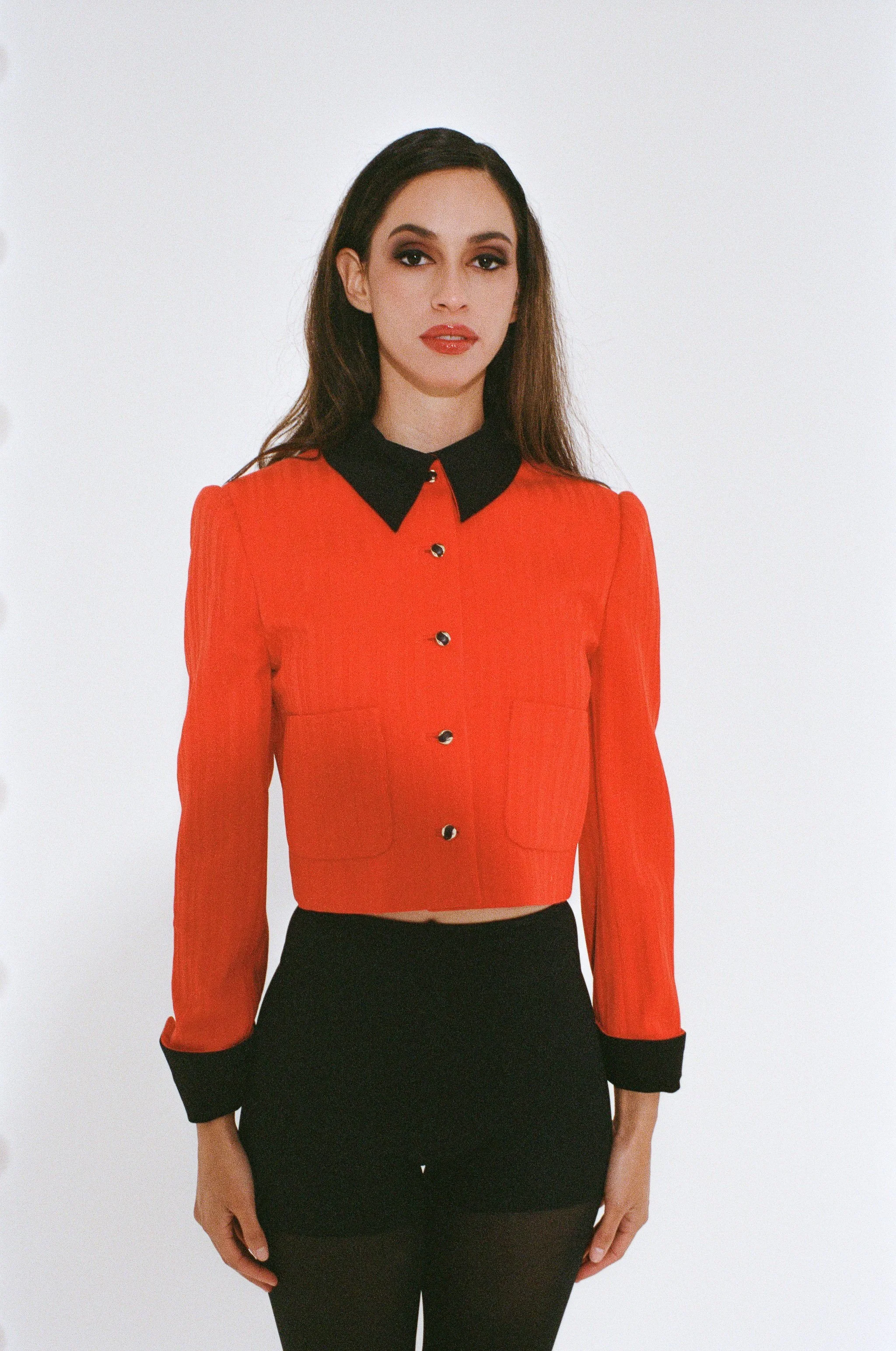 Fashion model wearing a red jacket with a black collar and buttons, black pants, and standing against a white studio background.