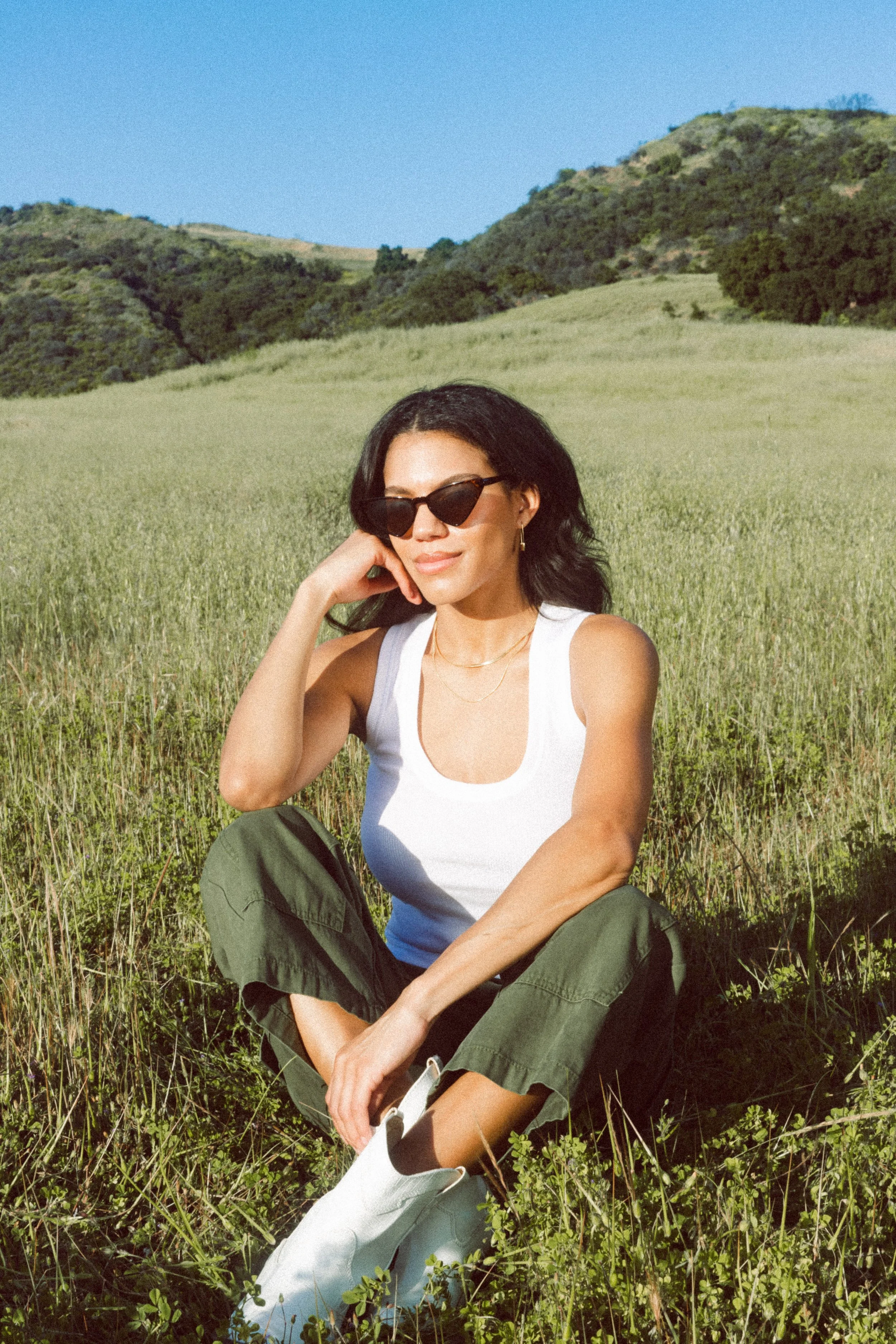 A model sitting in a grassy field with rolling hills in the background, wearing sunglasses, a white tank top, green cargo pants, and white boots.
