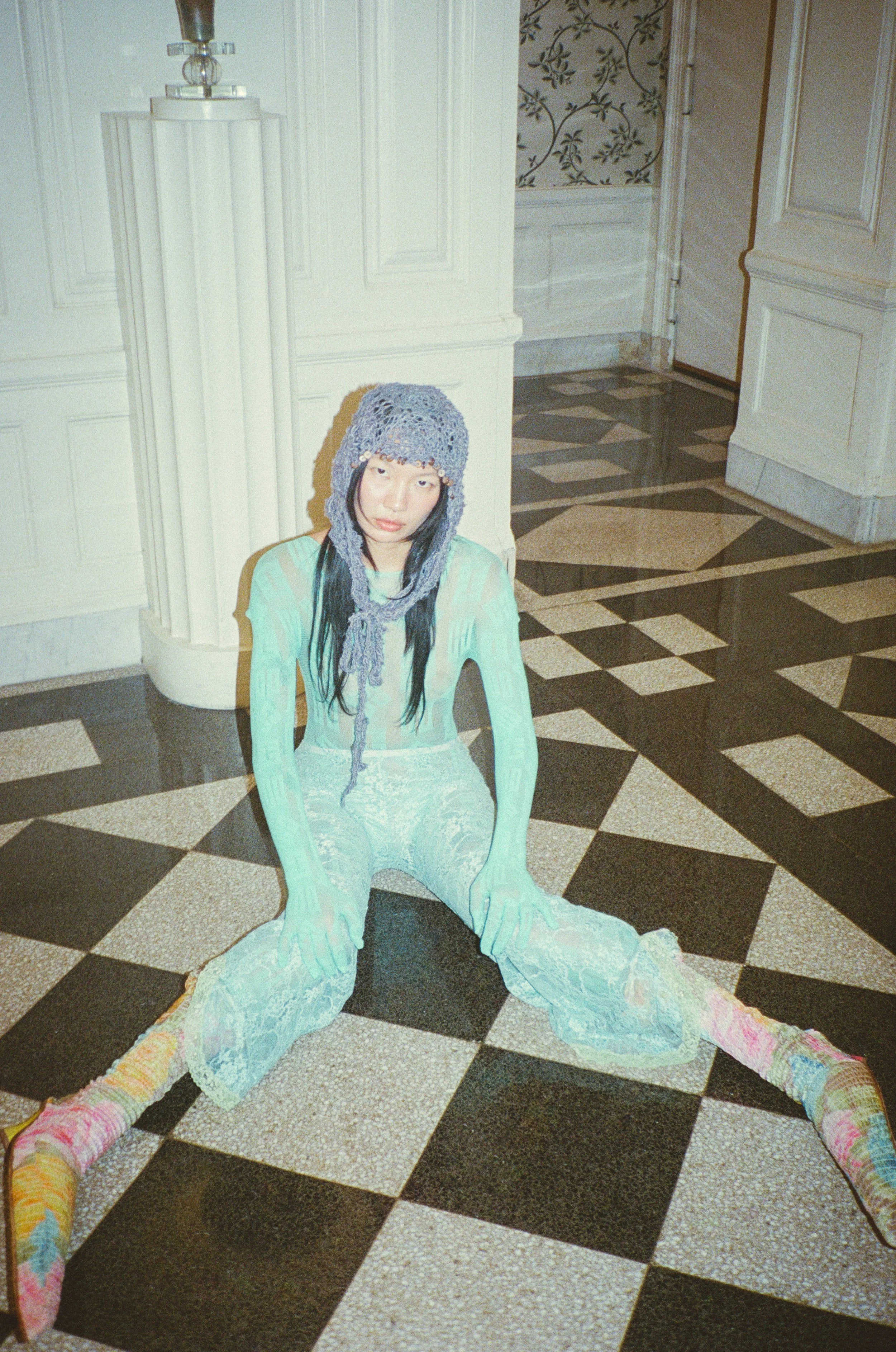 Fashion model sitting on a black-and-white tiled floor with legs outstretched, wearing pastel-colored pants and socks in an ornate interior.