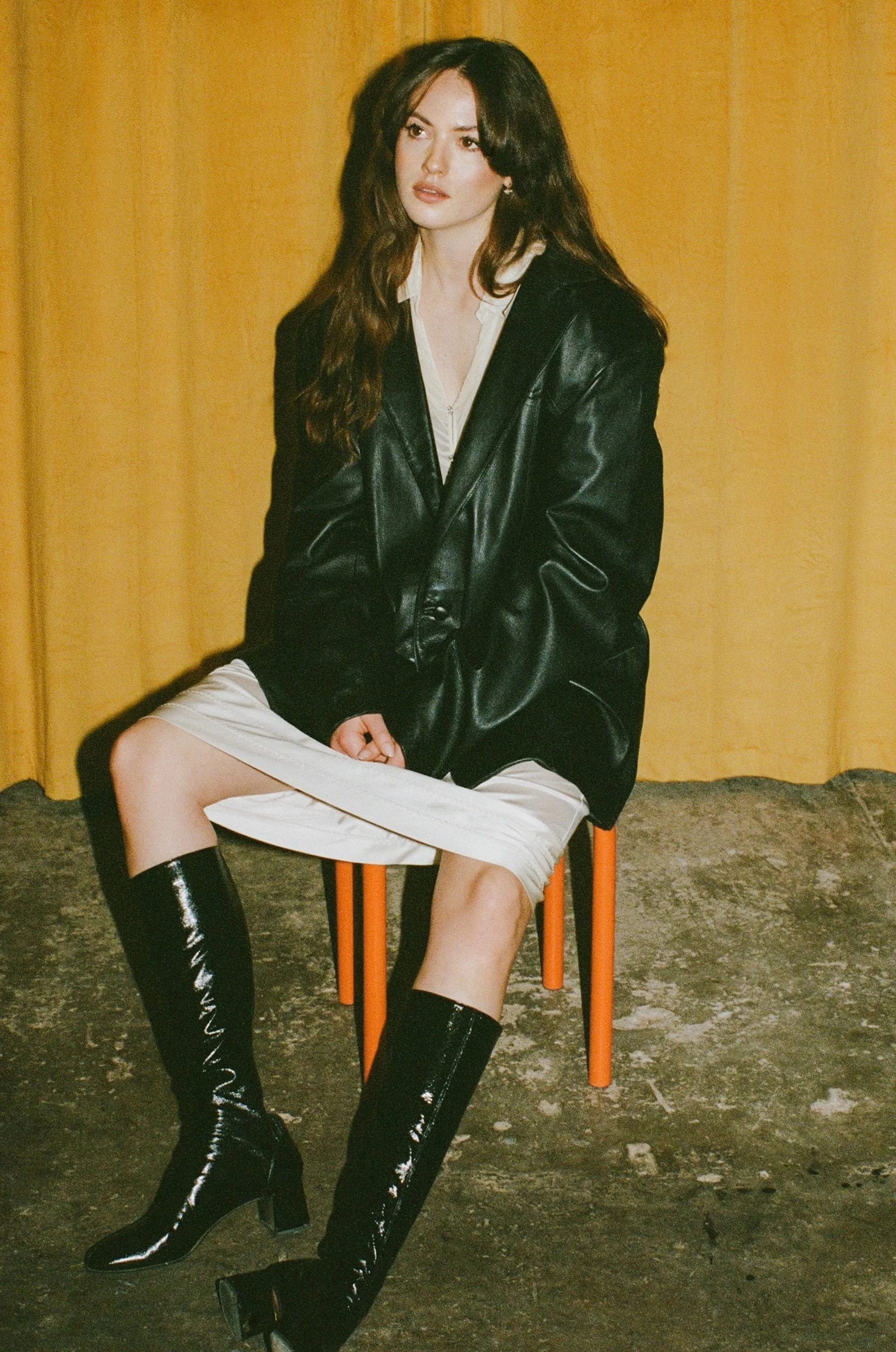 Fashion model with long wavy brown hair wearing a black leather jacket, white dress, and black knee-high boots, sitting on an orange chair in front of a yellow curtain.