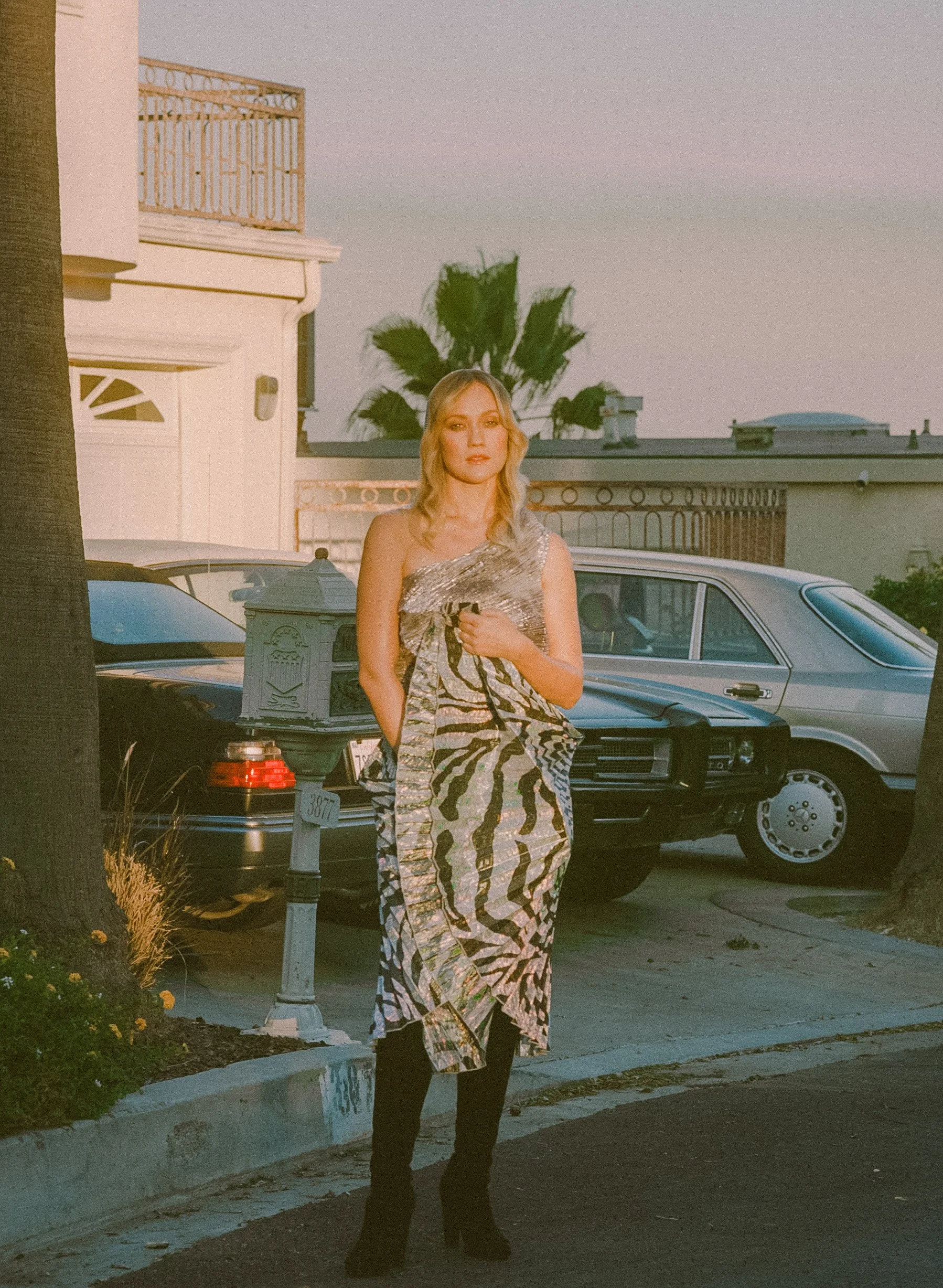Fashion model wearing a shimmering patterned long-sleeve dress, standing on a sidewalk near parked cars with palm trees and houses at sunset.