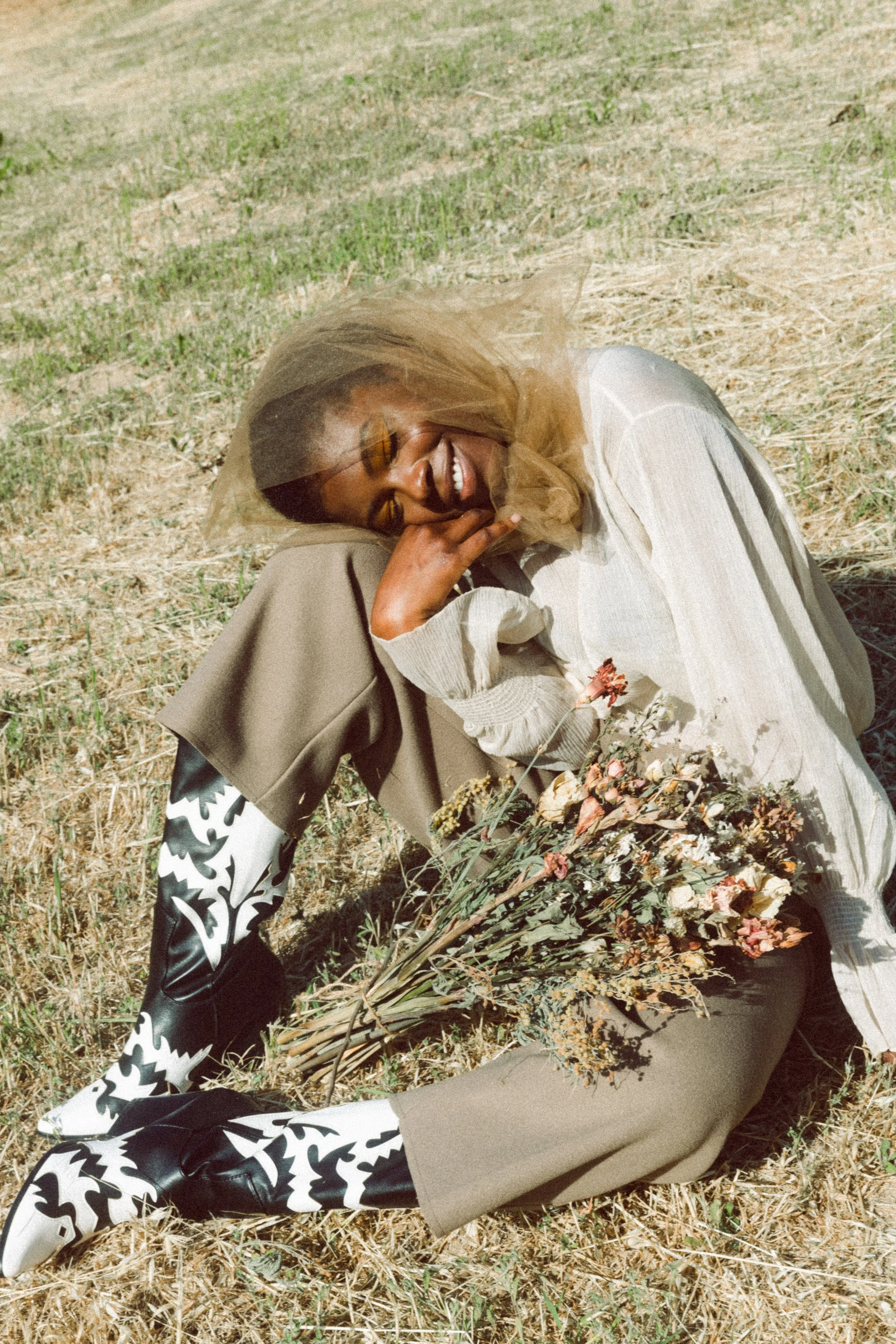 Fashion model with dyed hair lying on the ground outdoors, wearing a beige jacket with patterned sleeves, beige pants, black shoes, and a sheer beige headscarf while holding a branch of dried flowers.