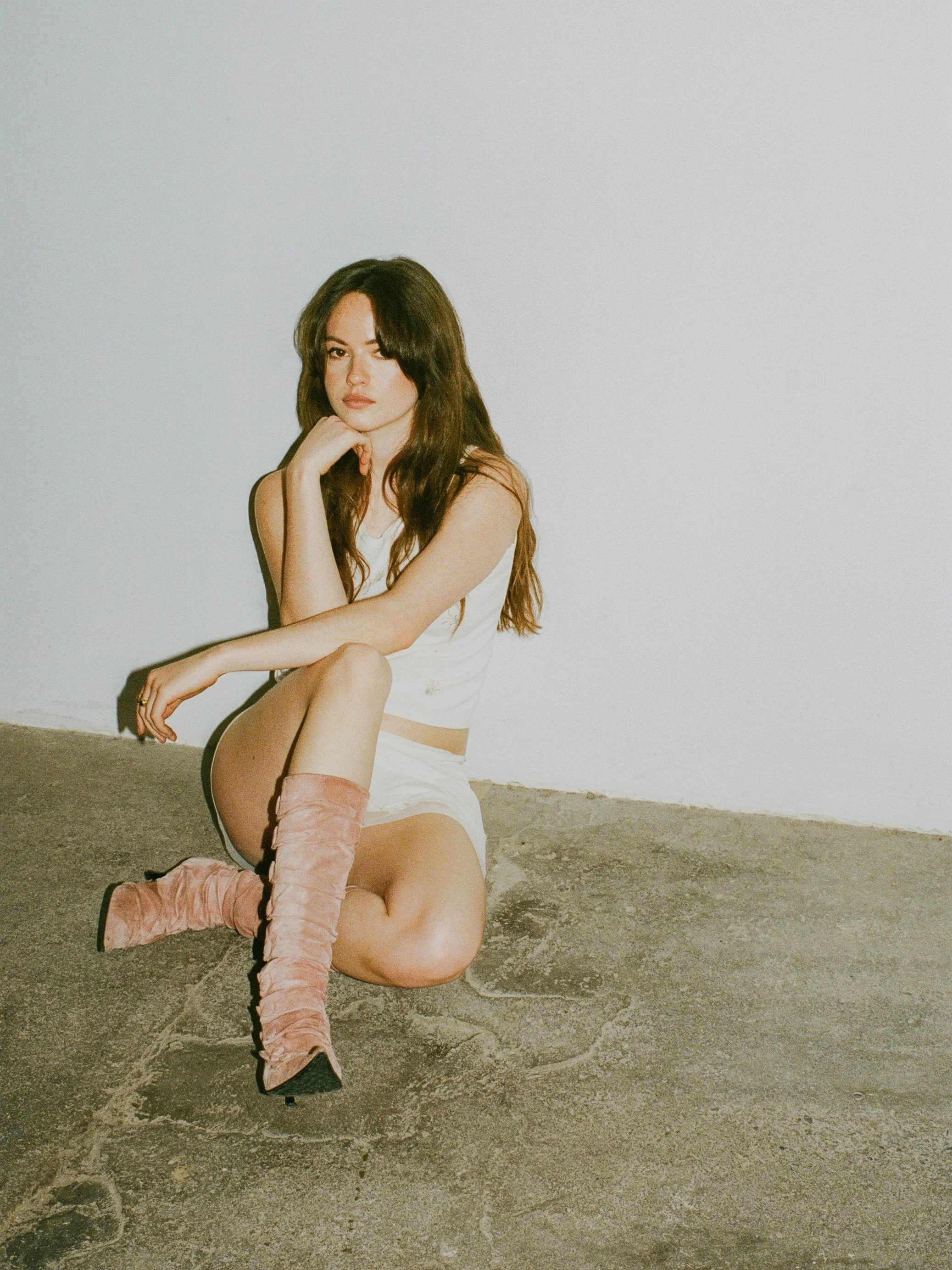 A woman with long brown hair sitting on a concrete ground against a plain white wall, wearing a white top, shorts, and pink high-heeled boots, with one knee raised and resting her chin on her hand.