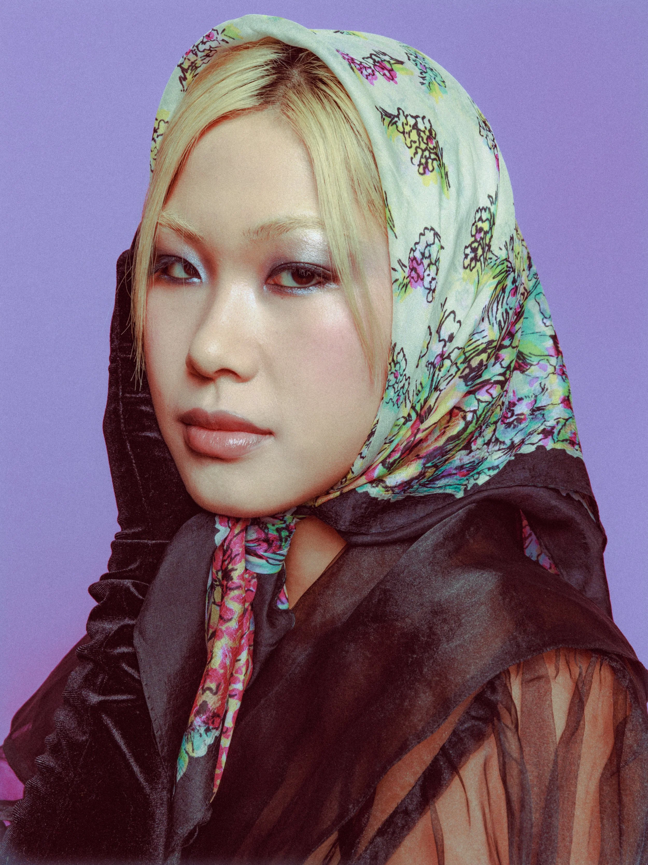 Editorial beauty portrait of a model wearing a colorful floral headscarf and dark sheer clothing, photographed against a soft lavender background.