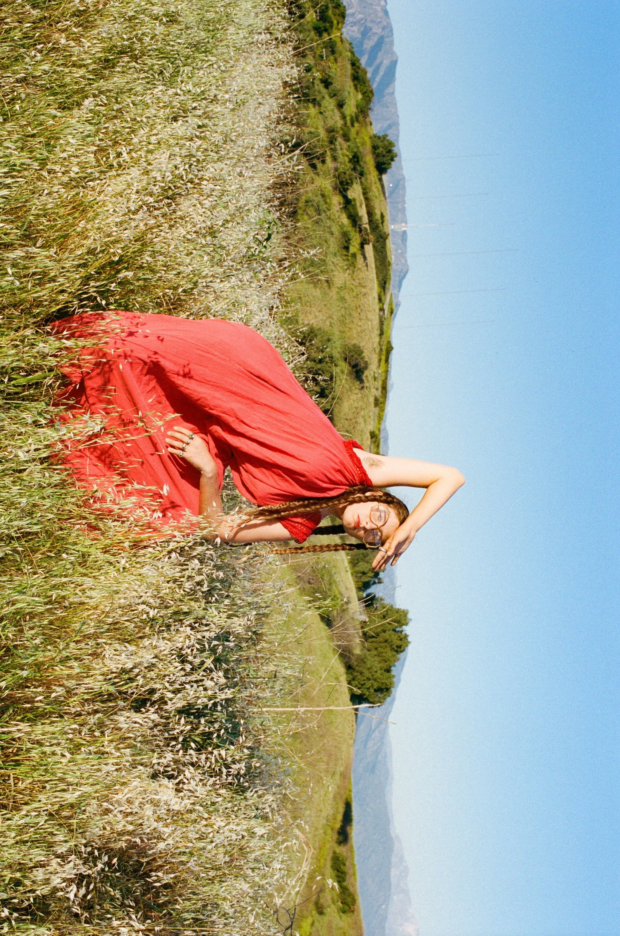 Fashion model in a red dress sitting in a field of tall grass and wildflowers with mountains in the background under a clear blue sky.