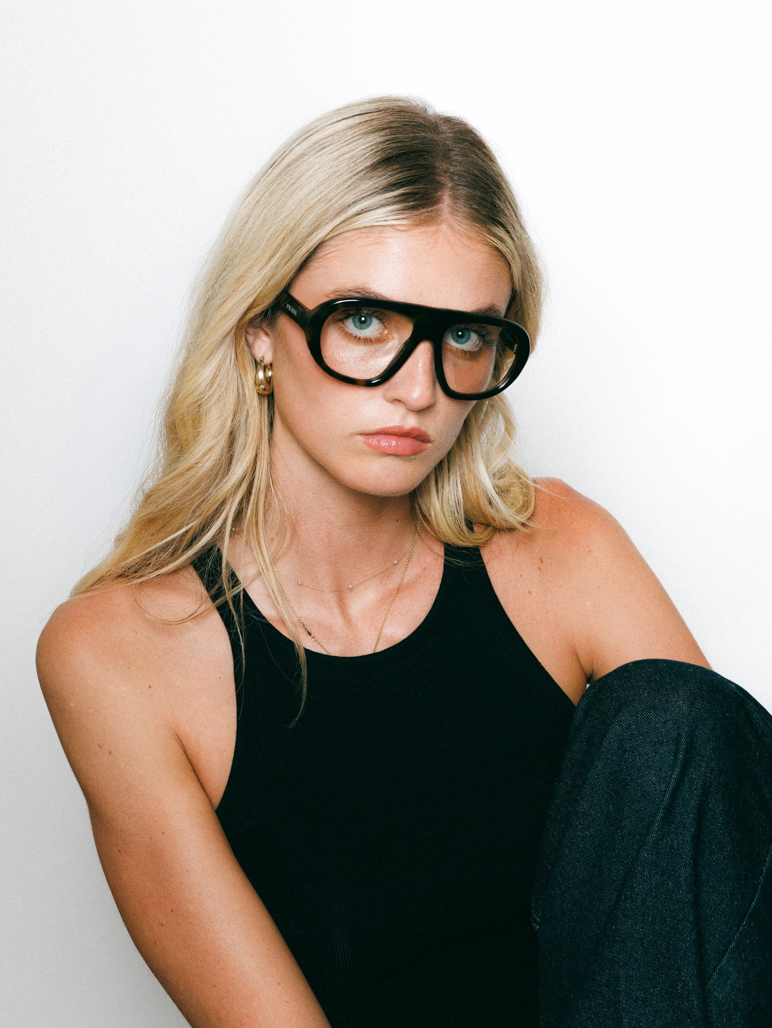Portrait of a blonde model wearing oversized black glasses and a black sleeveless top, seated against a clean white background.