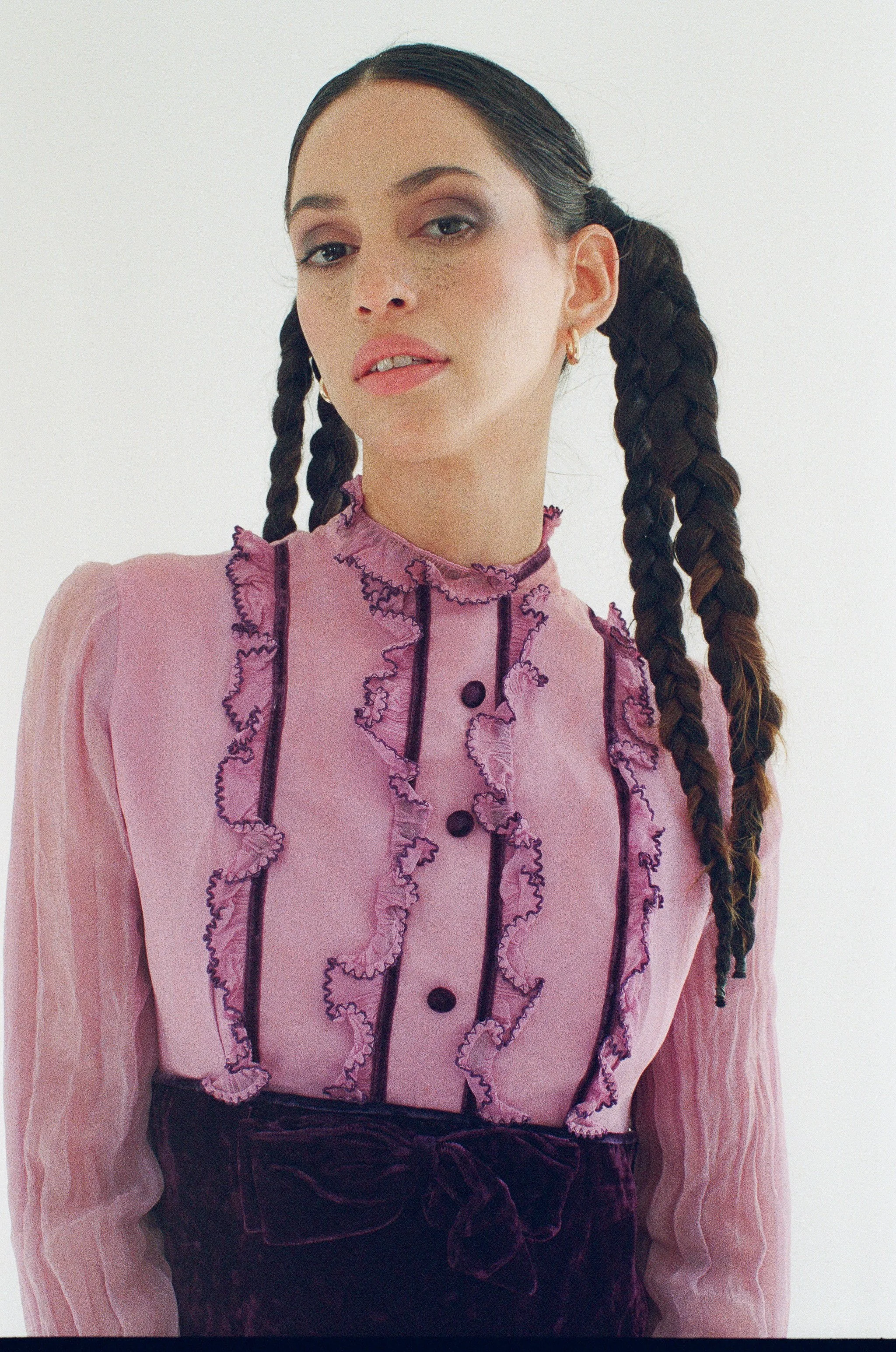 A model with dark braided hair, wearing a pink ruffled blouse with black accents, standing against a plain white background.