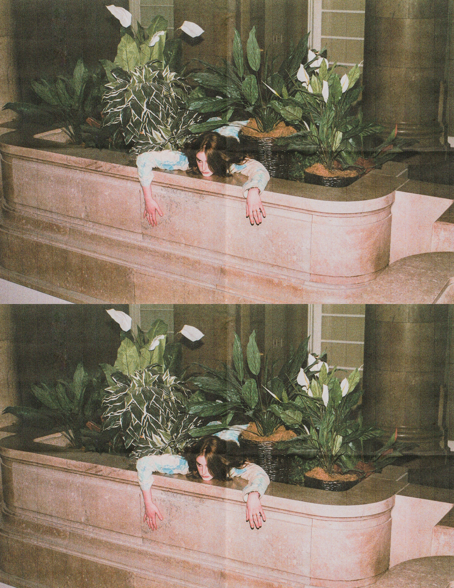 Fashion model leaning over a pink marble ledge with her head and arms hanging down, surrounded by large green houseplants indoors.