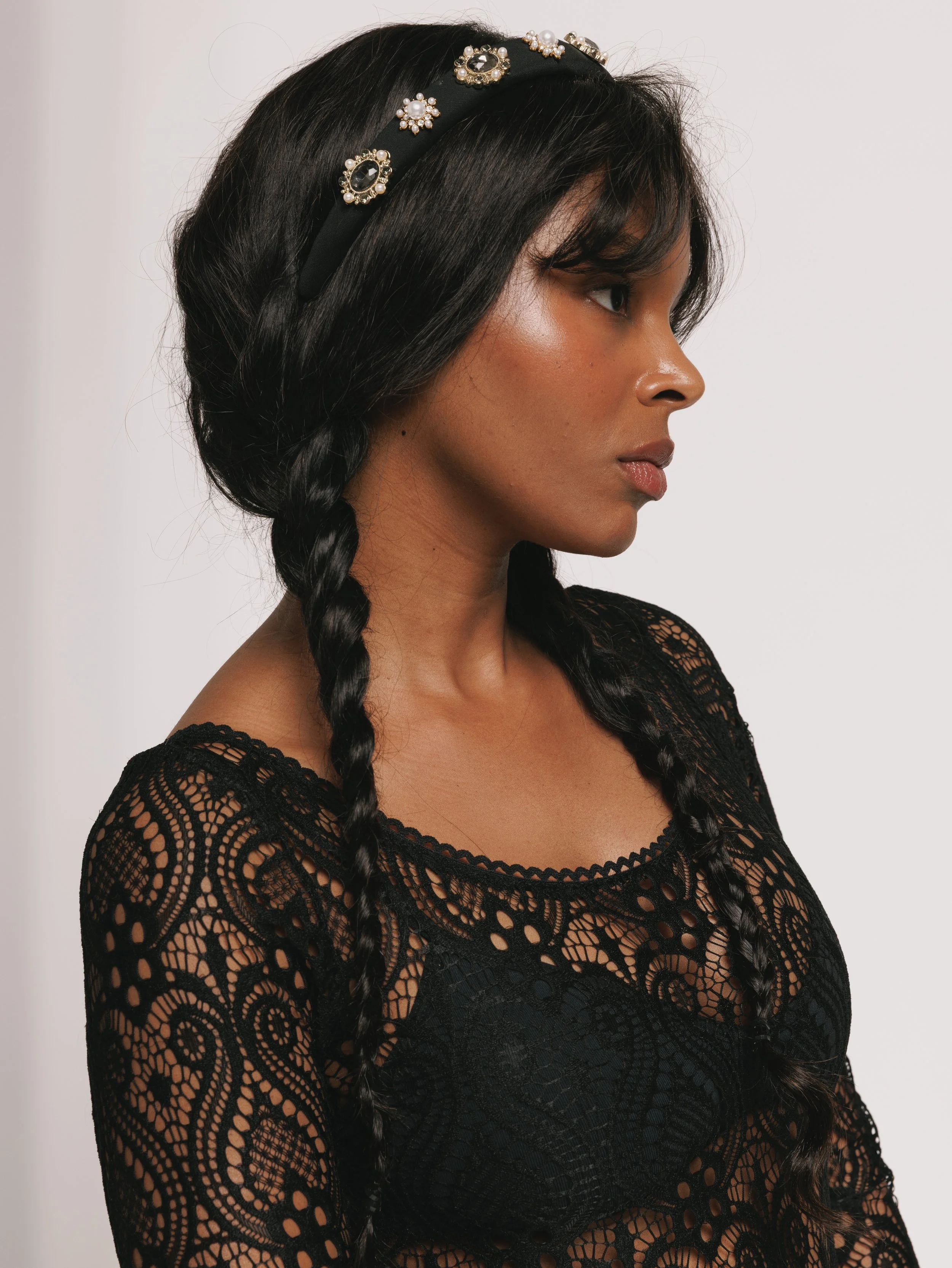Side profile of a fashion model with long black braided hair wearing a black lace top and a pearl- and jewel-embellished black headband.