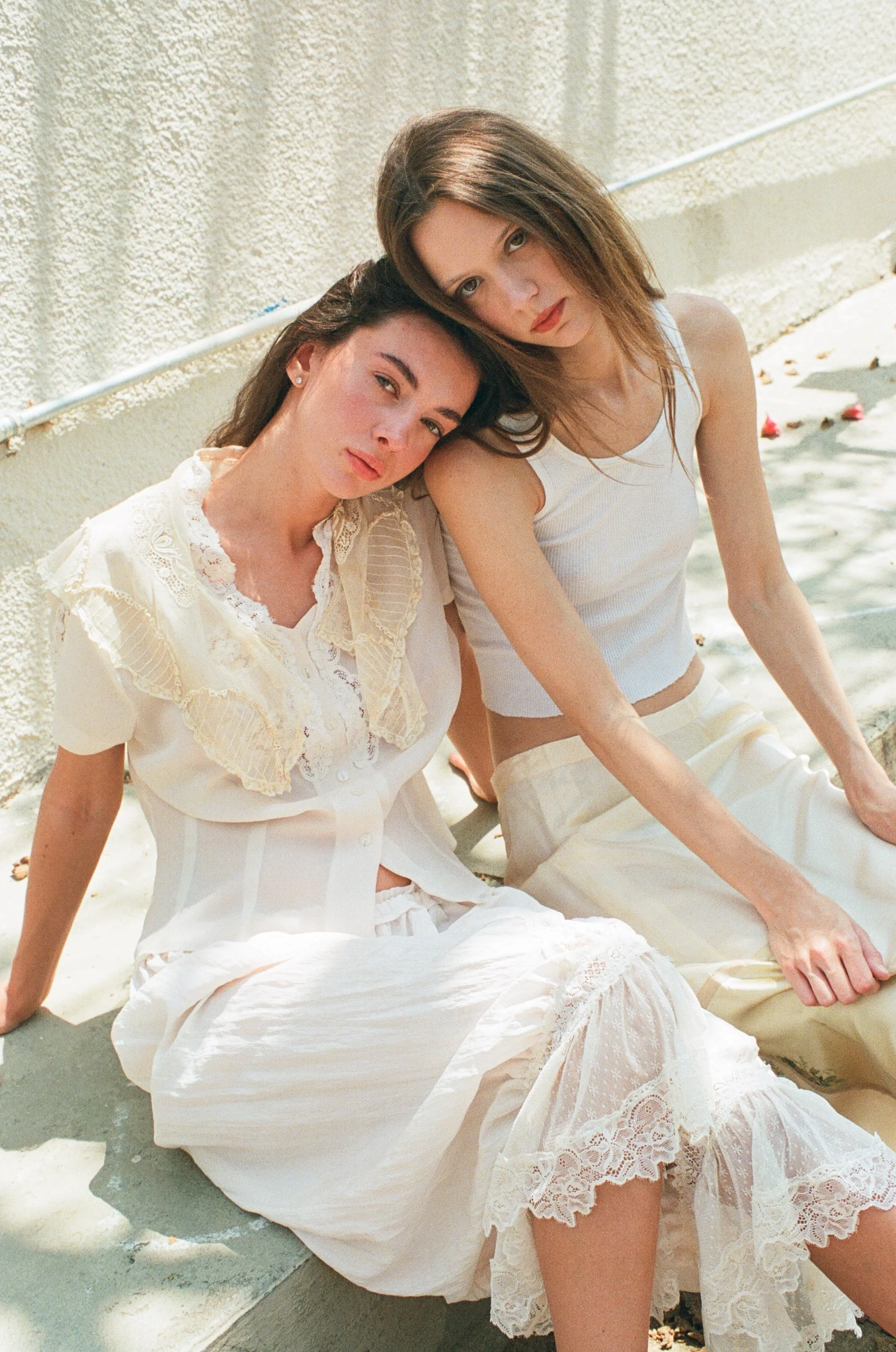 Two young fashion models sitting against a textured outdoor wall wearing light-colored vintage clothing and looking at the camera.