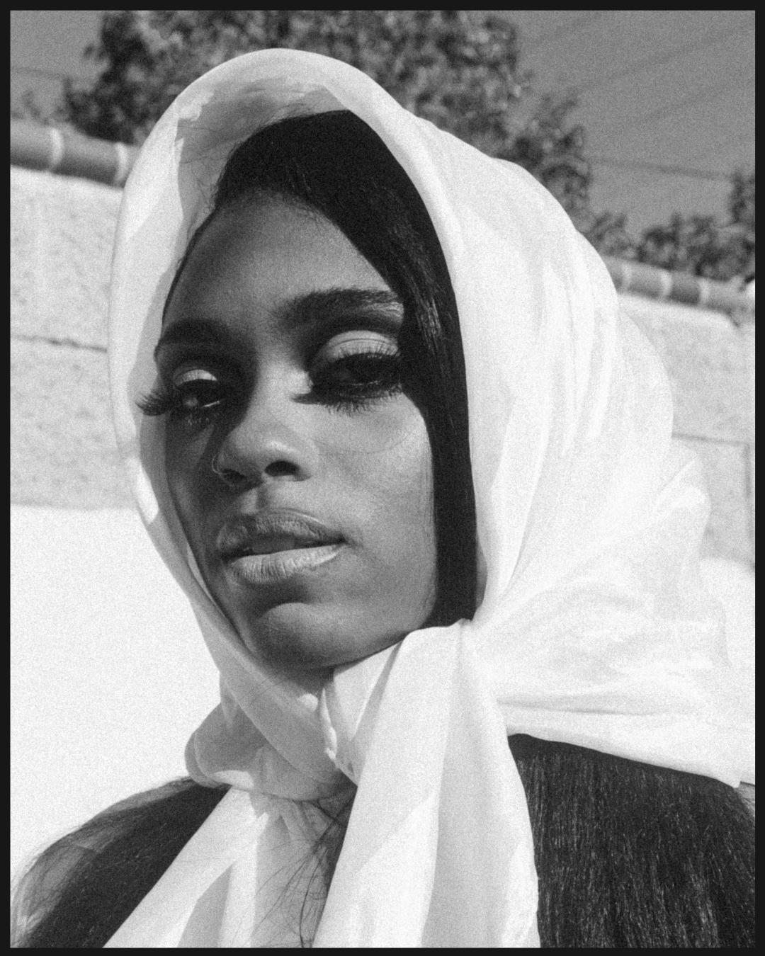 Black-and-white portrait of a woman wearing a light-colored headscarf, photographed outdoors in dramatic sunlight with a textured, film-like finish.