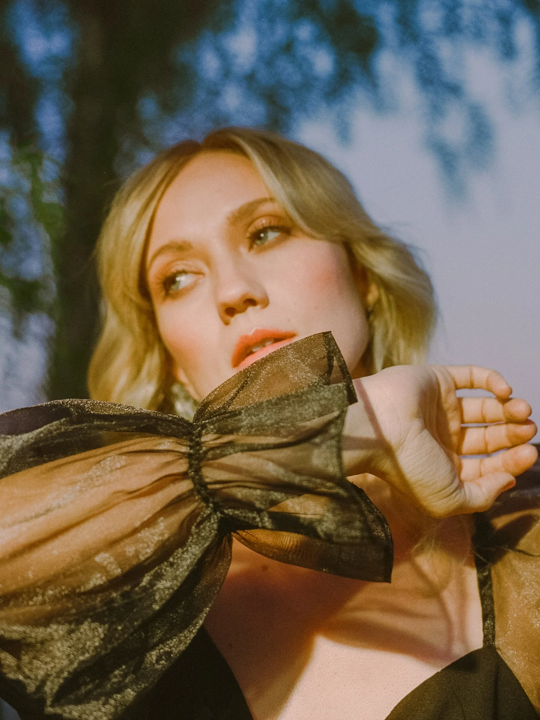 Close-up portrait of a fashion model with blonde hair holding black and gold sheer fabric near her face, with a softly blurred outdoor background.
