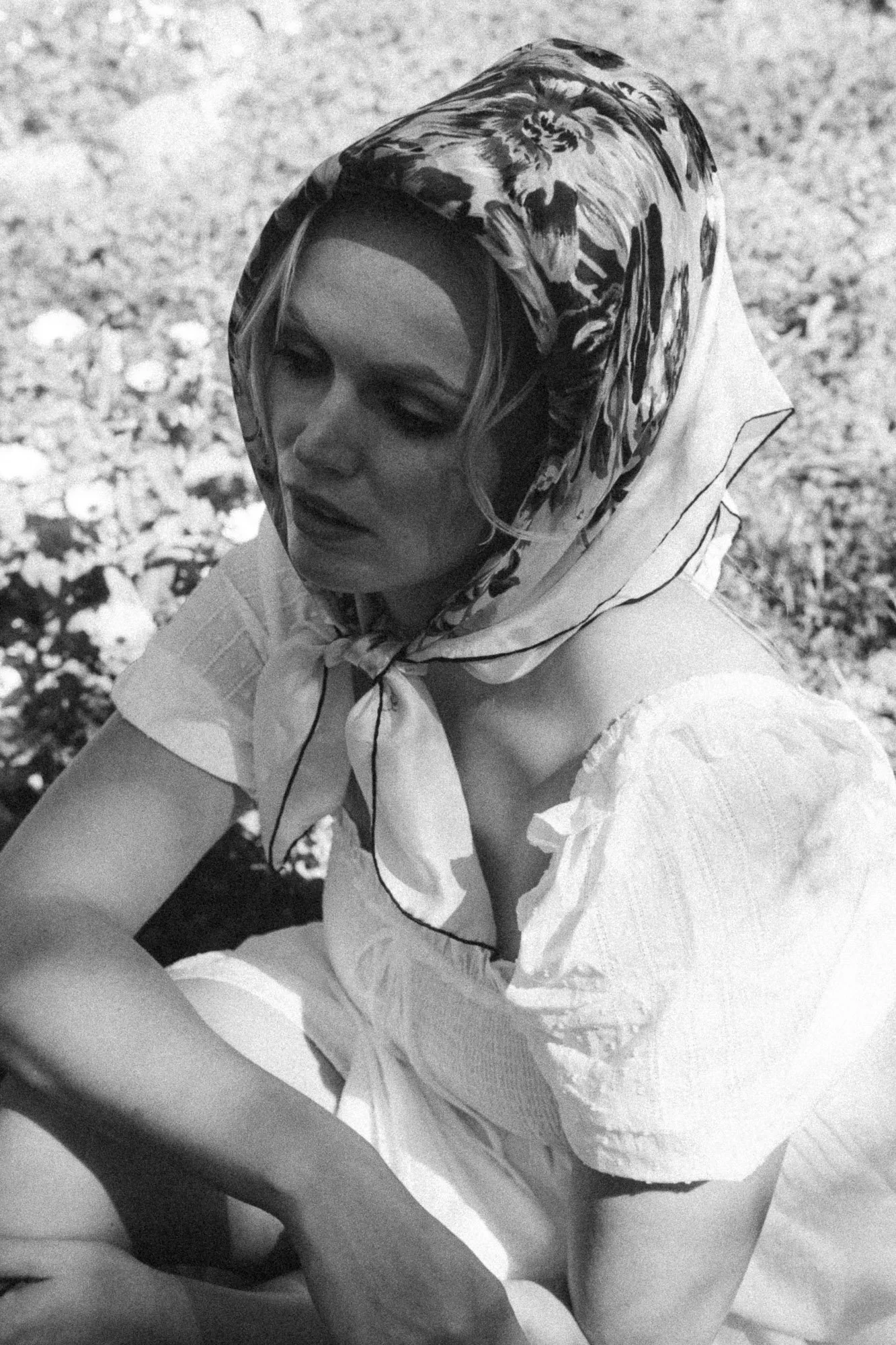 A woman with a headscarf looking down, wearing a white dress with puffed sleeves, sitting outdoors on gravel.