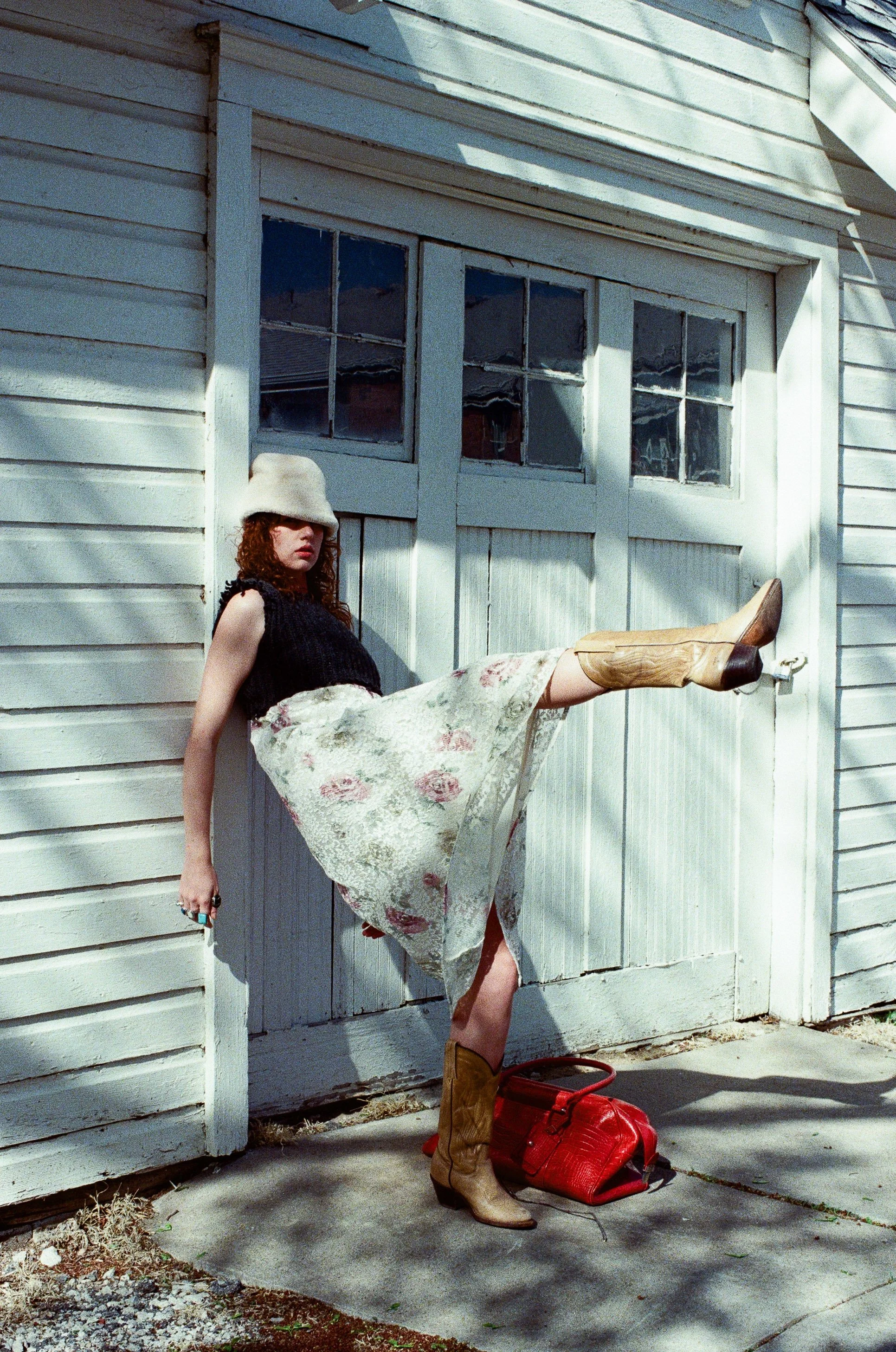 Fashion model with curly hair wearing a beige hat, black sweater, floral skirt, and cowboy boots, posing against a white garage door with a red bag on the ground.