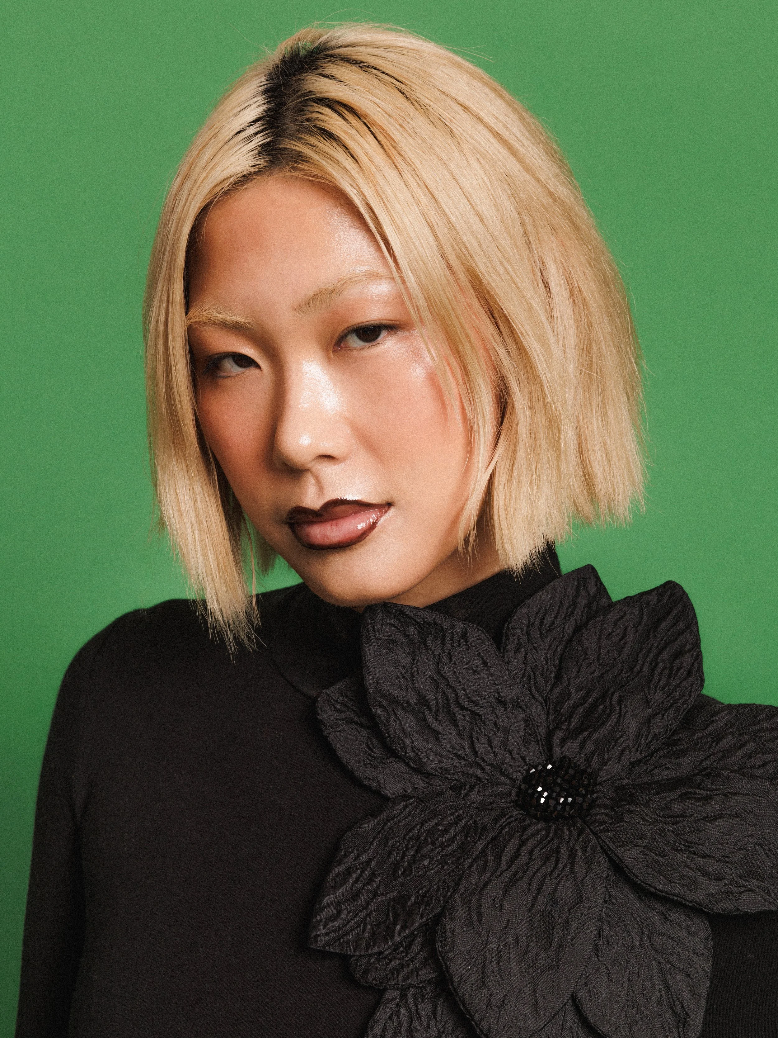 Editorial fashion portrait of a model wearing a black long-sleeve top with an oversized flower, photographed against a vibrant green studio background.