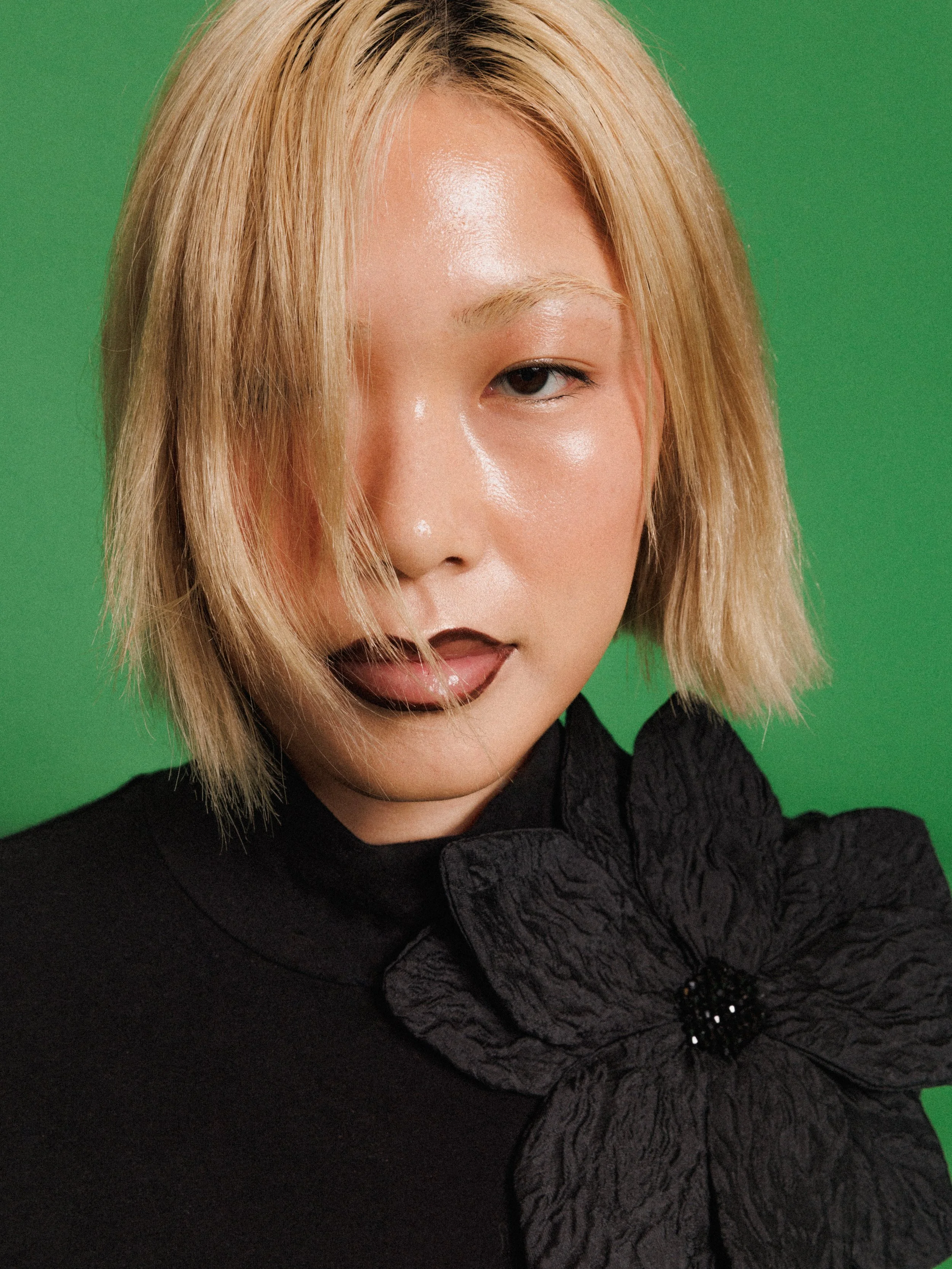 Editorial fashion portrait of a model wearing a black long-sleeve top with an oversized flower, photographed against a vibrant green studio background.