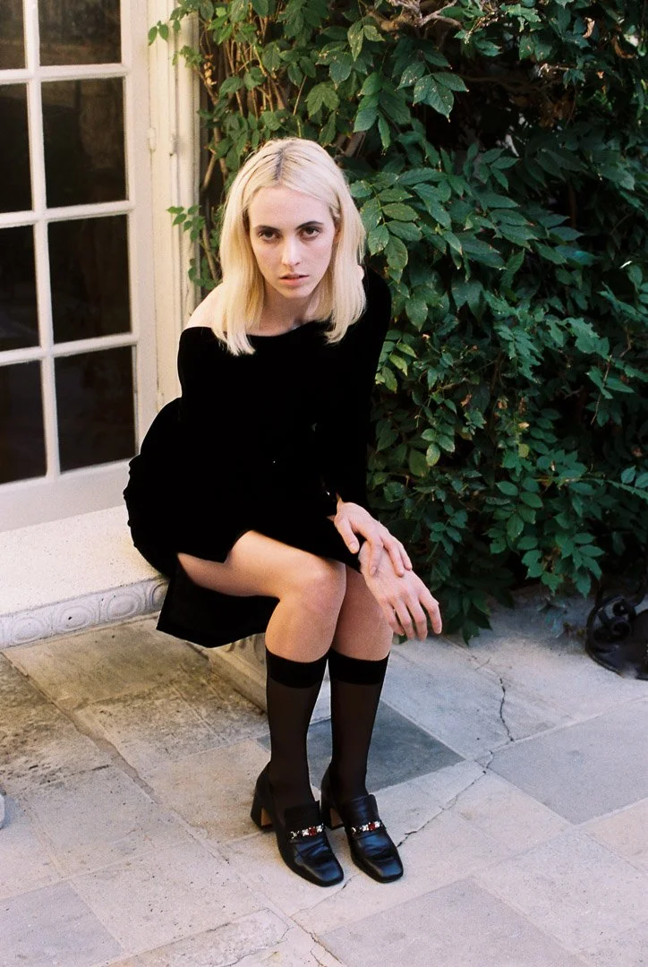 Fashion model with blonde hair wearing a black dress, black socks, and black shoes, sitting on a stone bench with greenery and a white door in the background.