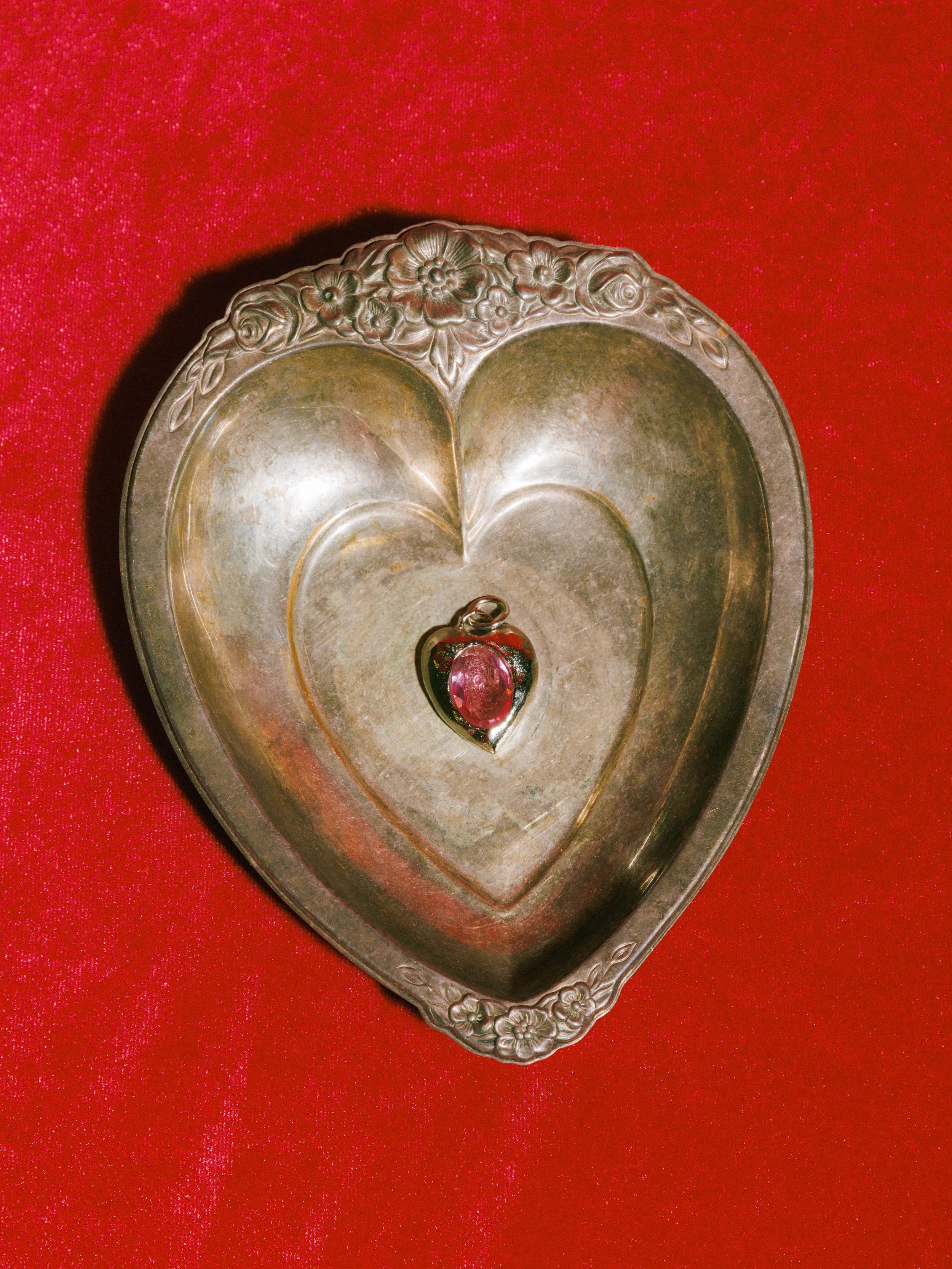 Silver heart-shaped tray with floral engravings holding a pink gemstone pendant on a red textured surface.