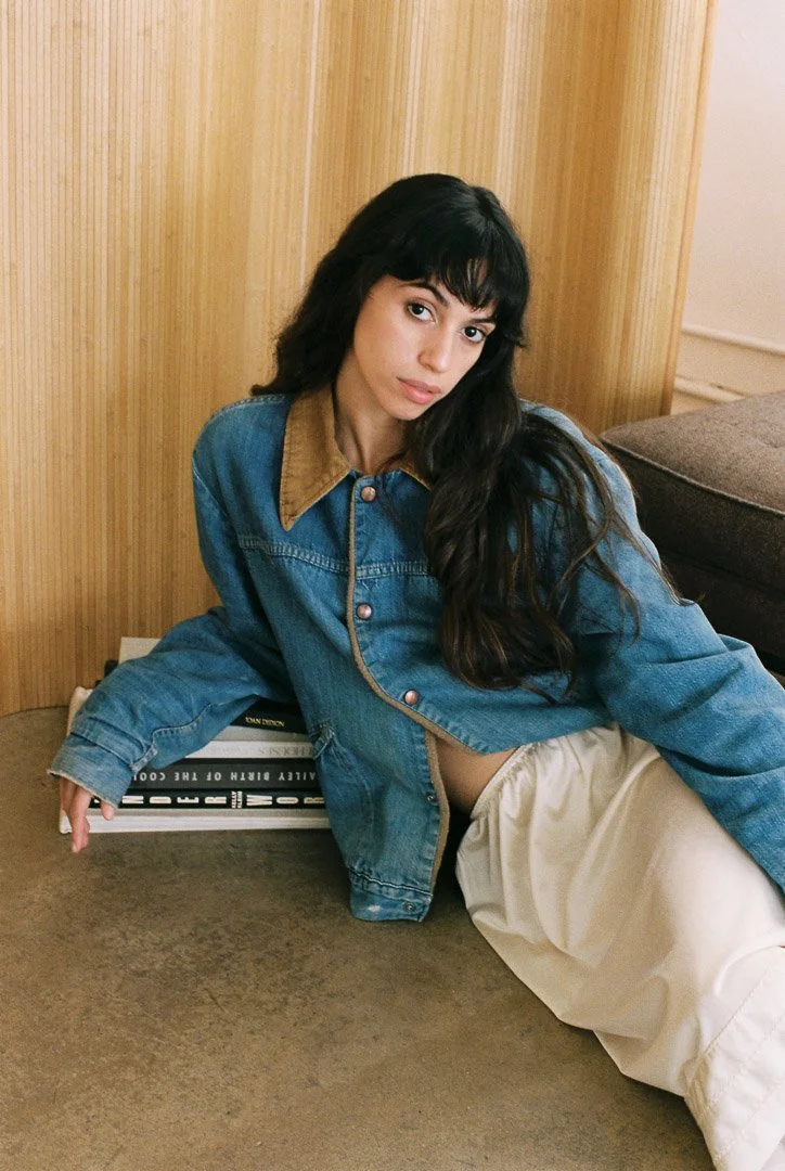 Editorial portrait of a fashion model wearing a blue denim jacket seated on the floor against a wooden wall.