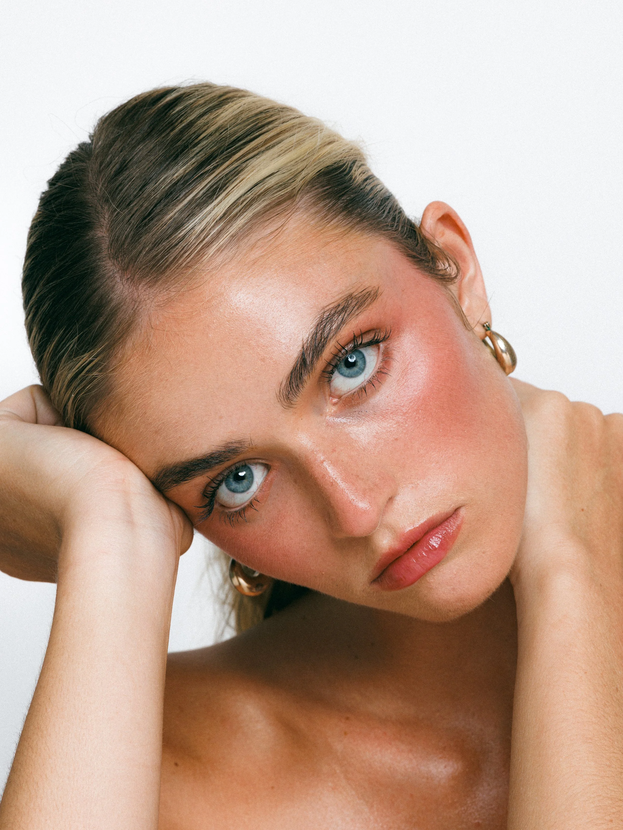 Close-up beauty portrait of a blonde model with glowing makeup, gold hoop earrings, and a sleek ponytail against a white background.