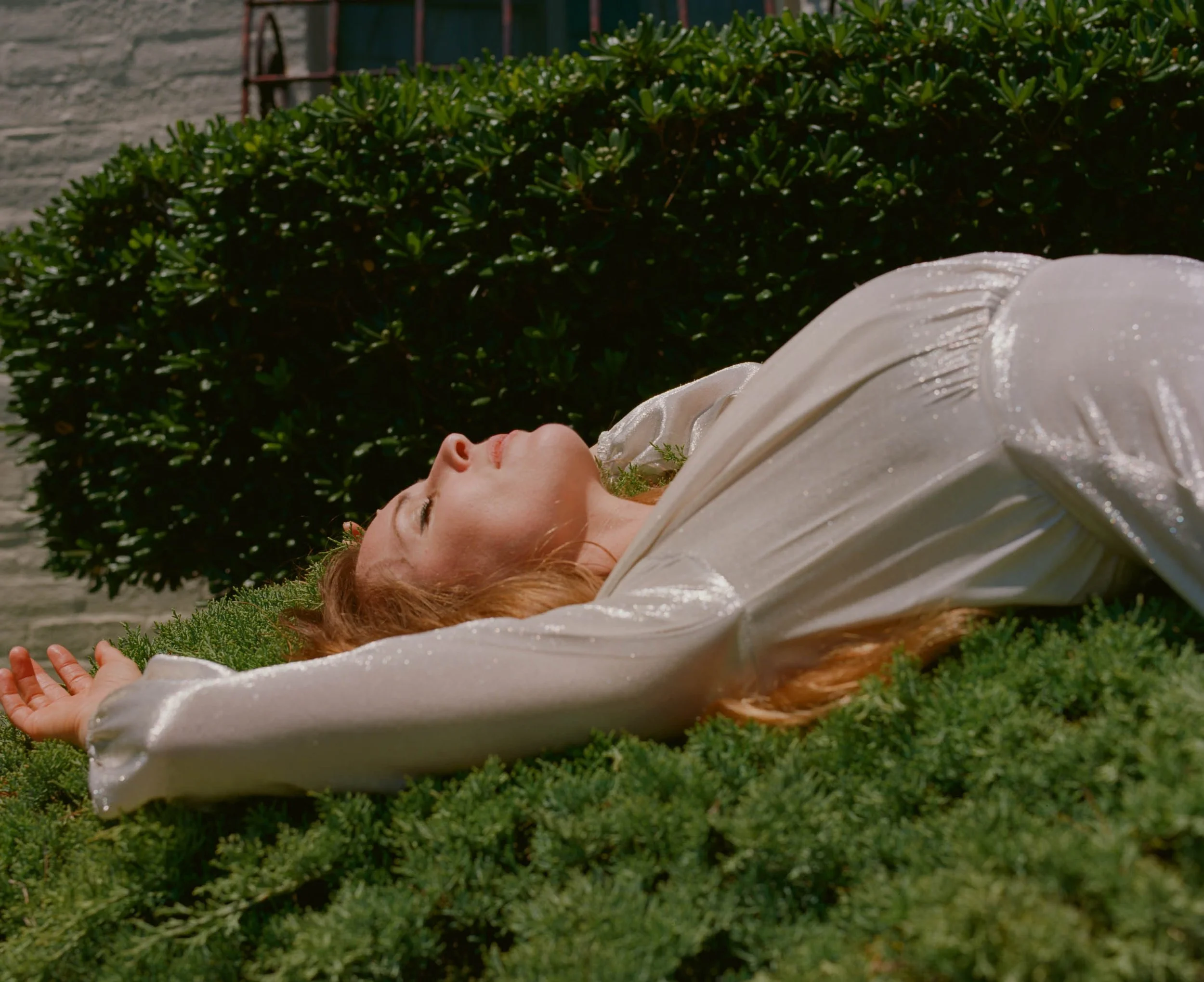 Fashion model lying on grass with arms outstretched, wearing a cream-colored dress with a brick wall and greenery in the background.