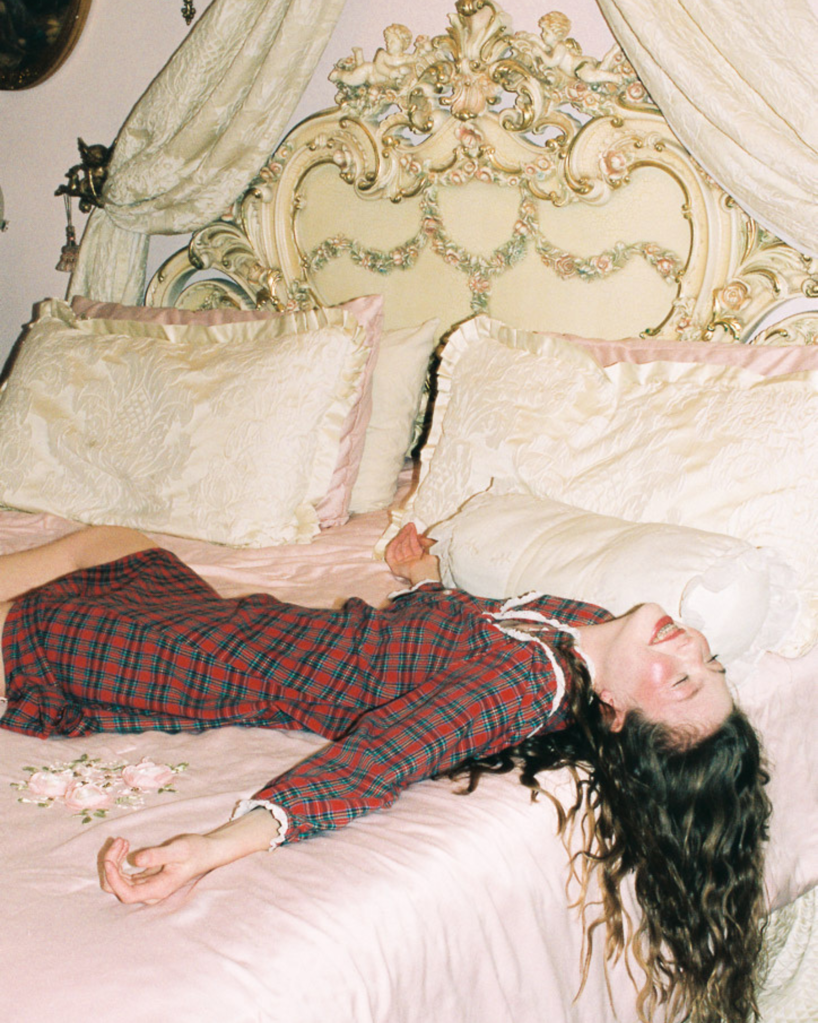 Editorial fashion photograph of a model in a red plaid dress reclining across an ornate vintage bed with pink satin bedding and romantic décor.