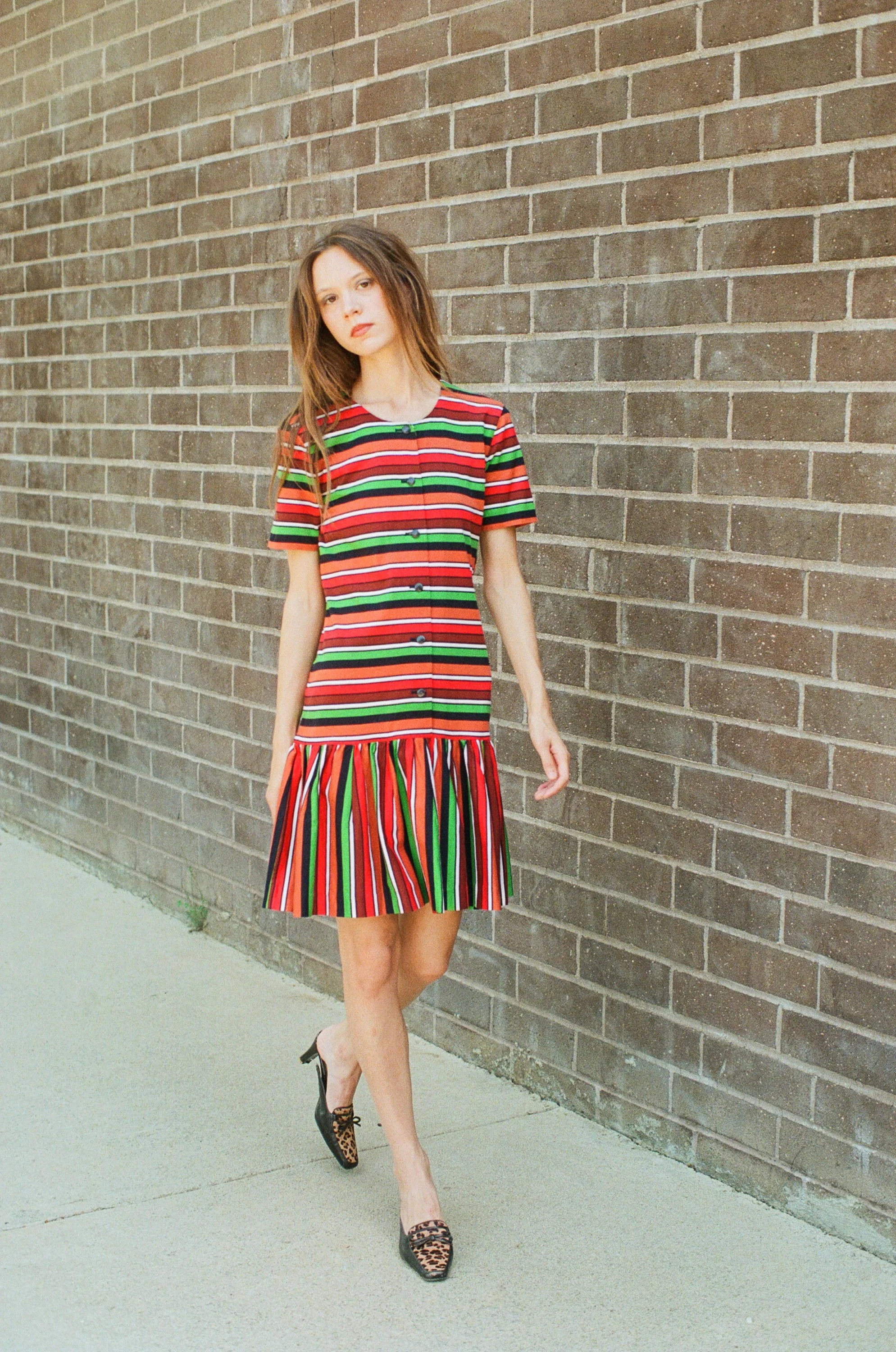 Fashion model standing against a brick wall outdoors wearing a colorful striped dress and leopard-print shoes.