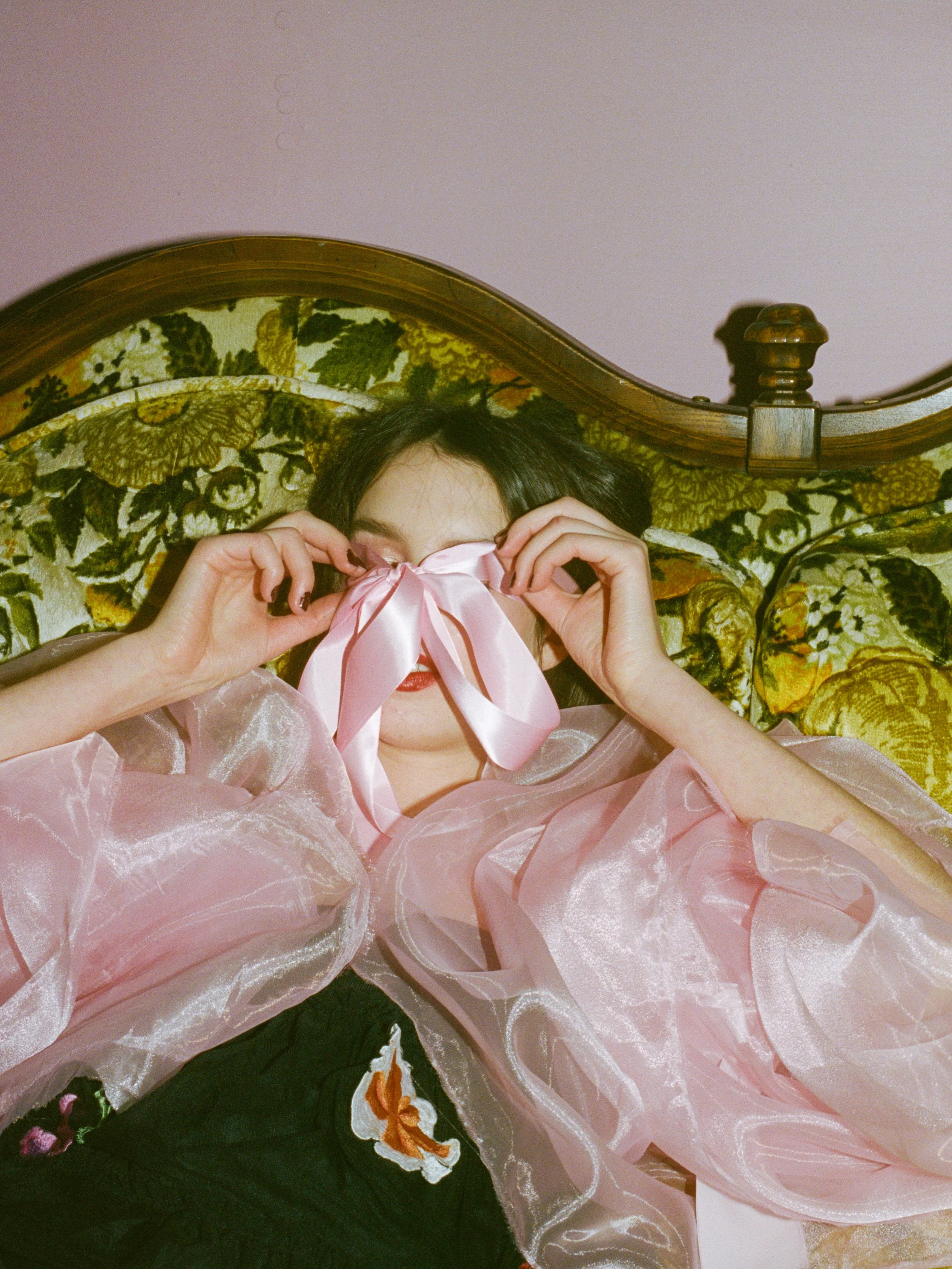 Fashion model with dark hair and red lipstick lying in bed, holding a pink satin ribbon over her eyes, with a vintage floral headboard in the background.
