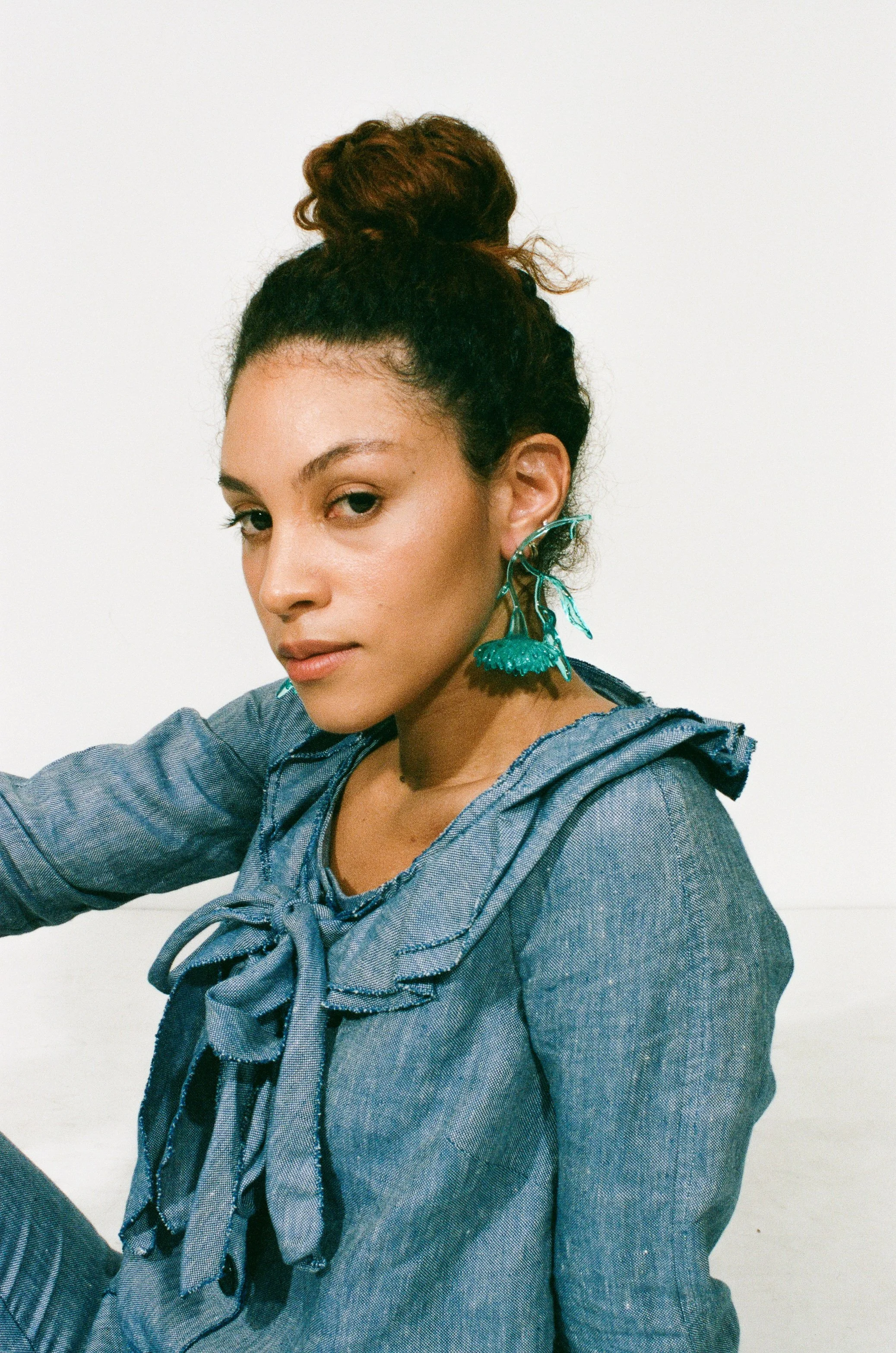 Portrait of a fashion model with curly hair in a high bun wearing large teal earrings and a denim shirt with a bow-tied collar against a white background.