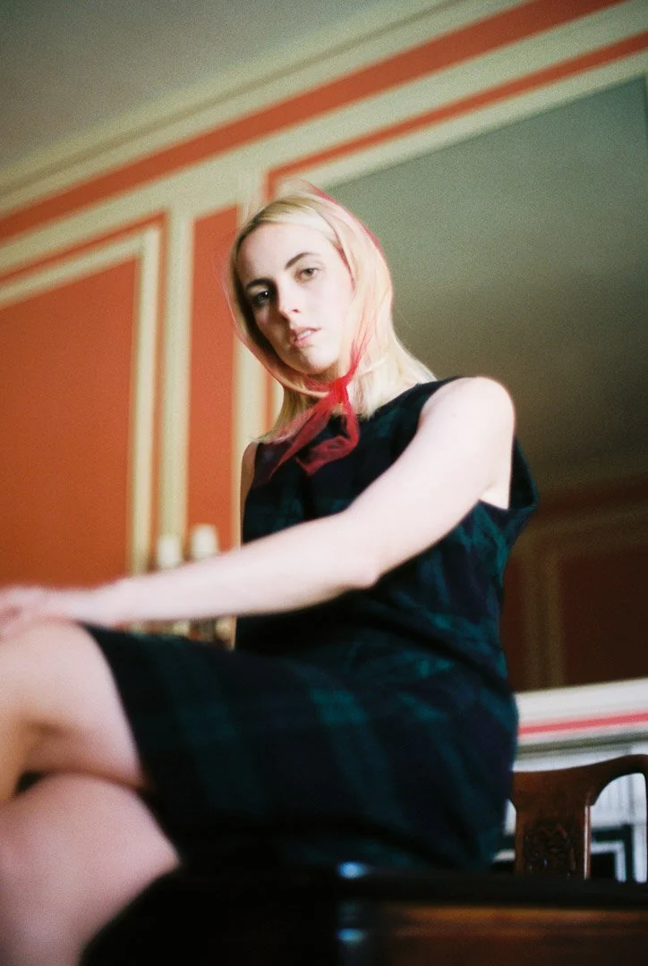 Fashion model with blonde hair wearing a black sleeveless dress and a red scarf, sitting on a chair in a room with red and white walls.