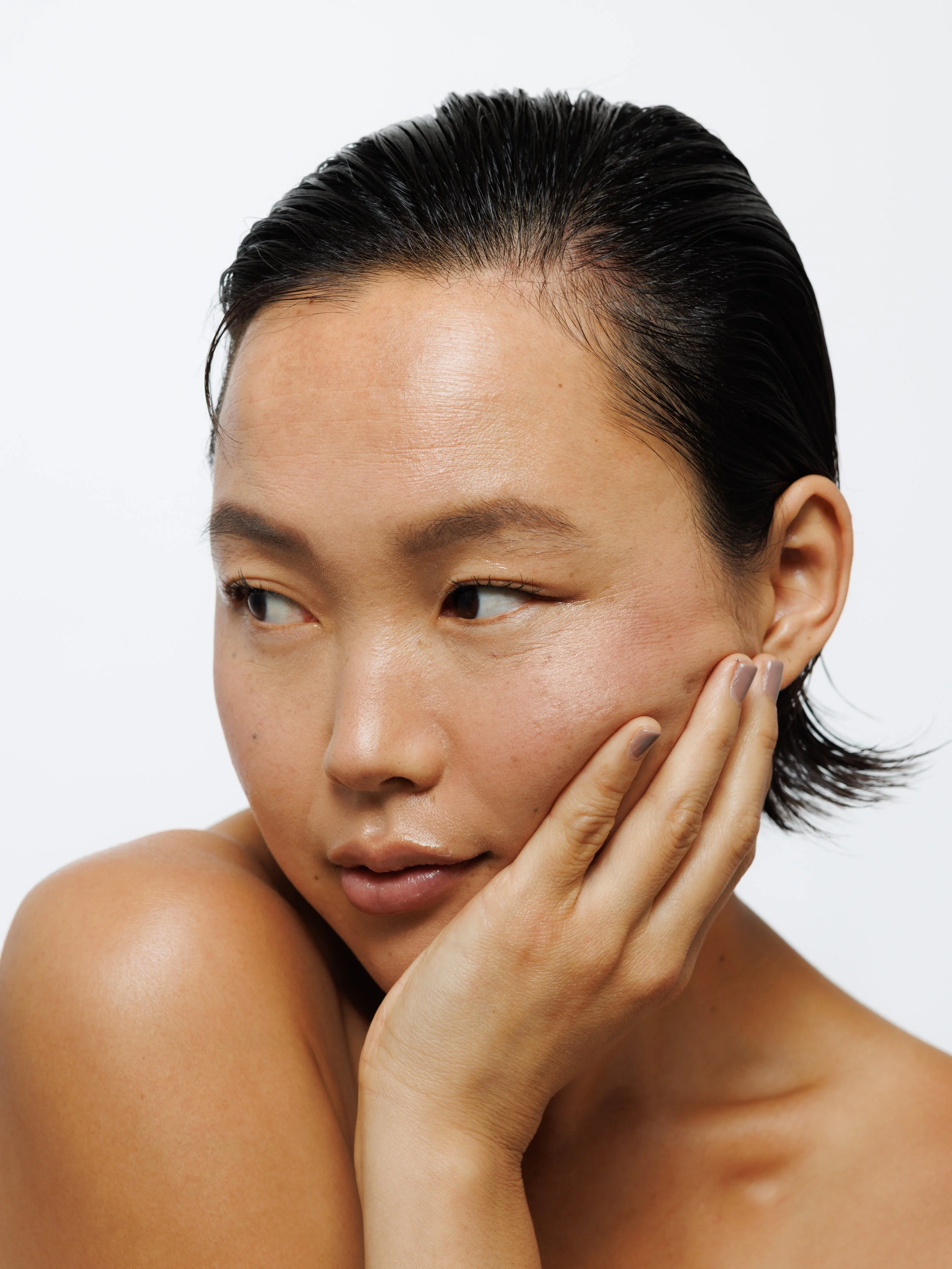 Close-up portrait of a fashion model with short black hair, holding her hand near her face with a relaxed expression.