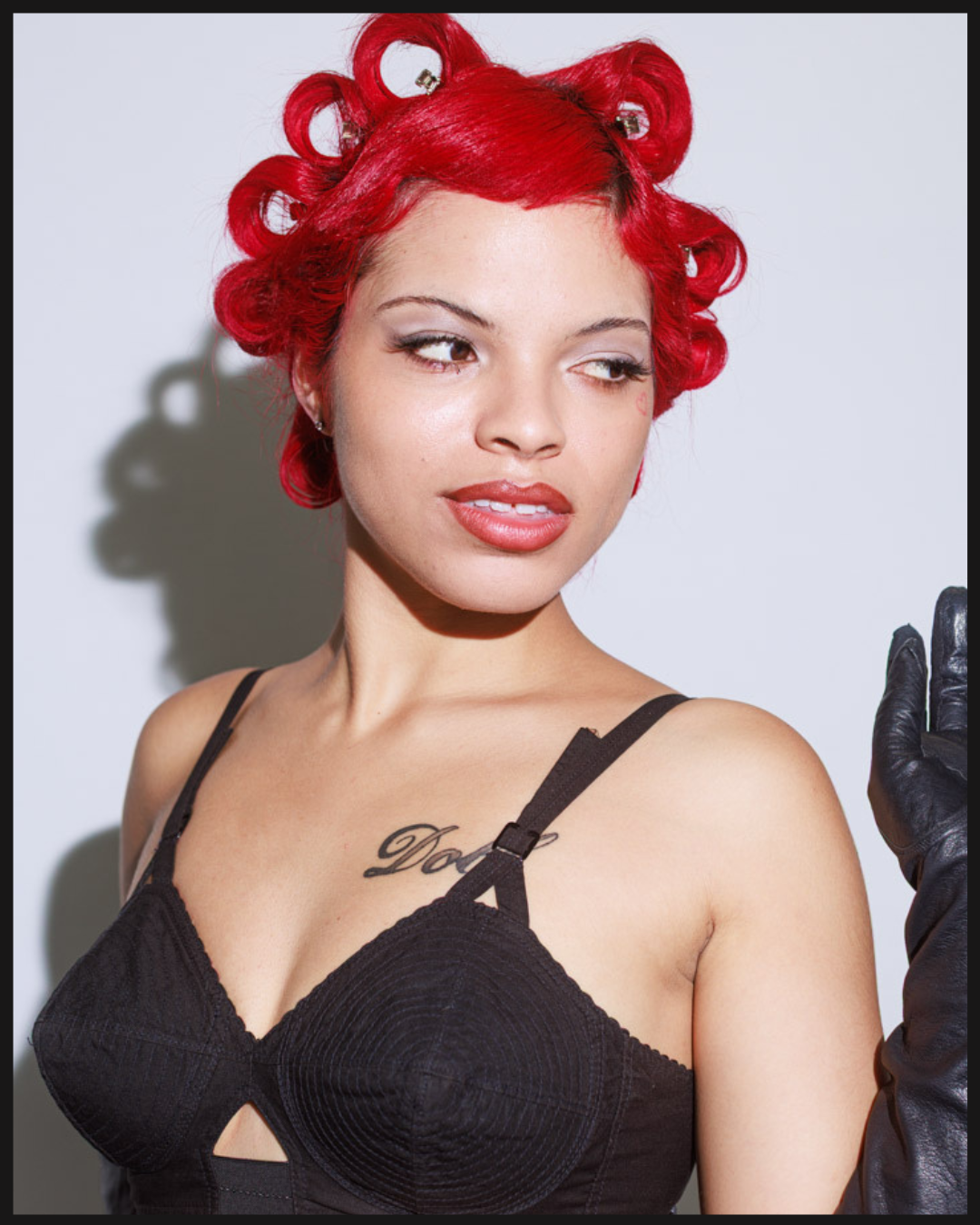 Portrait of a woman with bright red pin curls wearing a black top, photographed with direct flash against a light gray background.