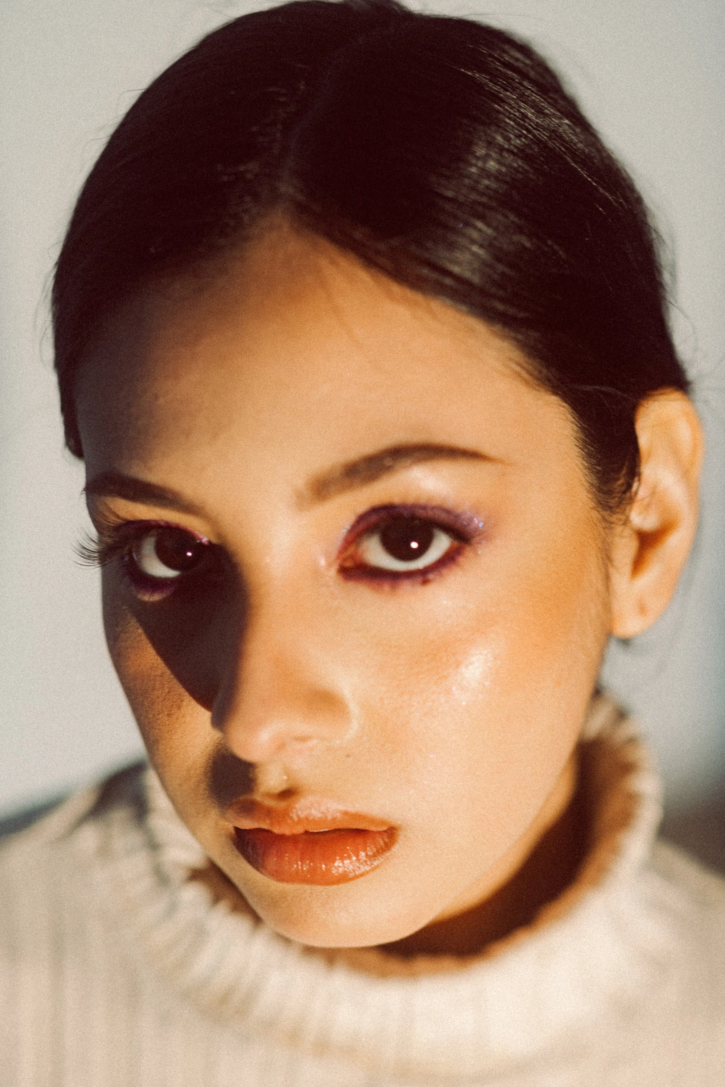 Close-up of a woman's face with dark hair, purple eyeshadow, and peach lipstick, wearing a white fluffy garment.