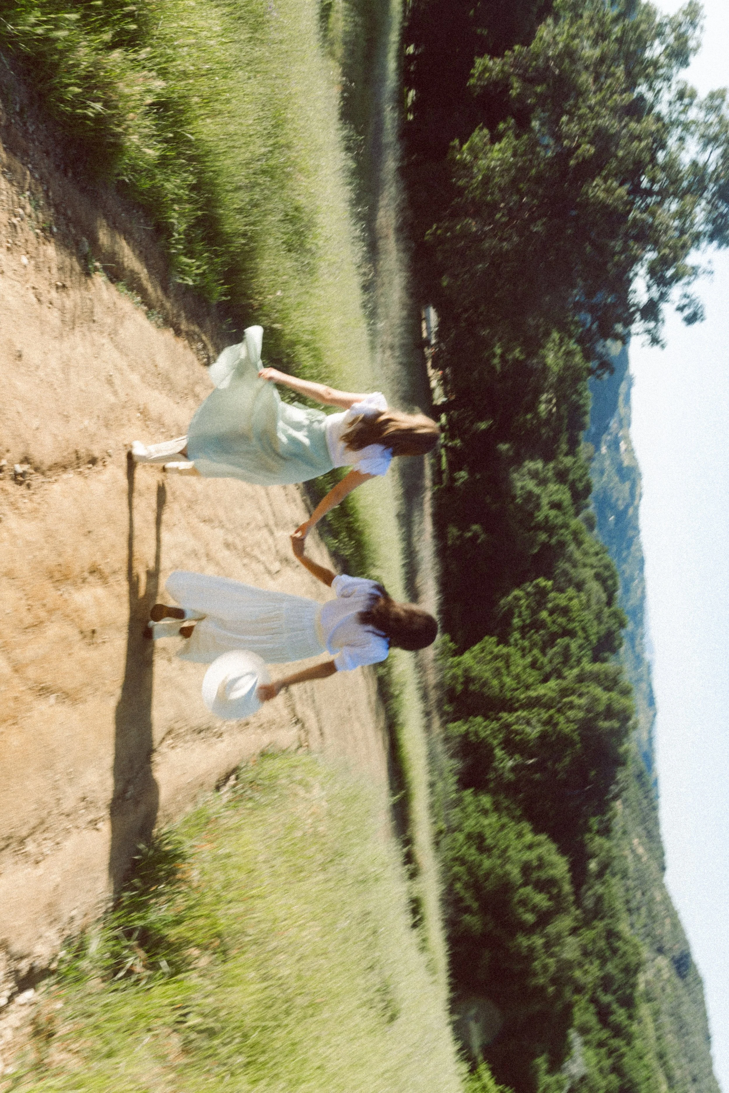 Two fashion models in white dresses and wide-brimmed hats walking along a dirt path through a green landscape with trees and mountains in the background.