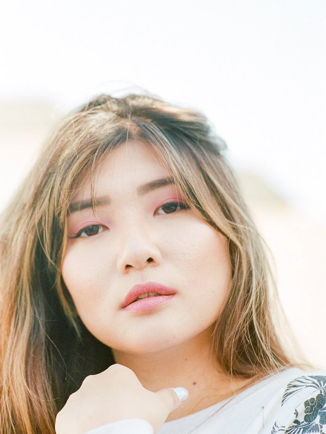A close-up photo of a young woman with long, light brown hair, makeup, and a white top with a black pattern. She is looking into the camera with a neutral expression.
