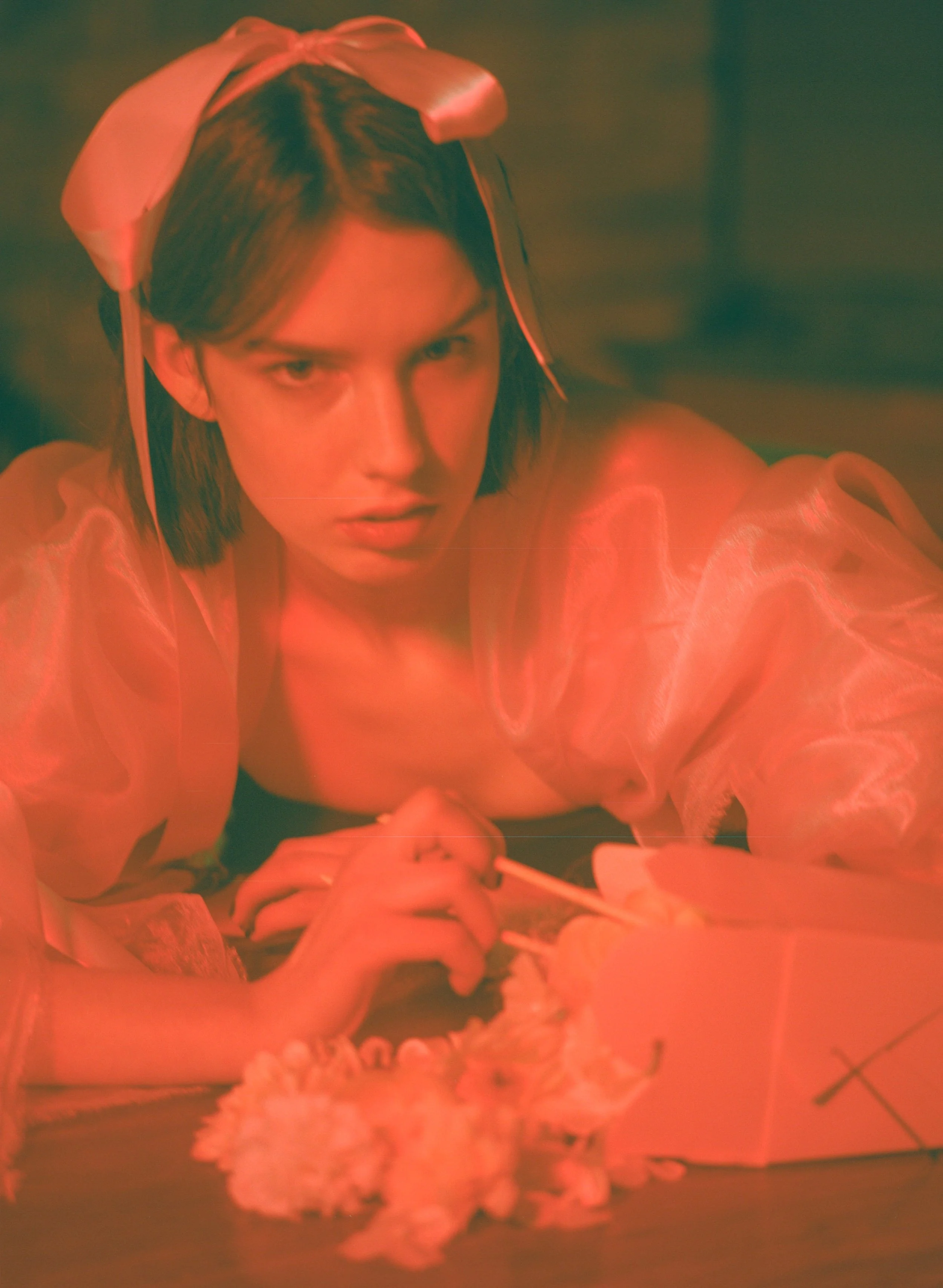 Fashion model with short dark hair wearing a pink bow headband, sitting at a table with flowers in warm, reddish light.