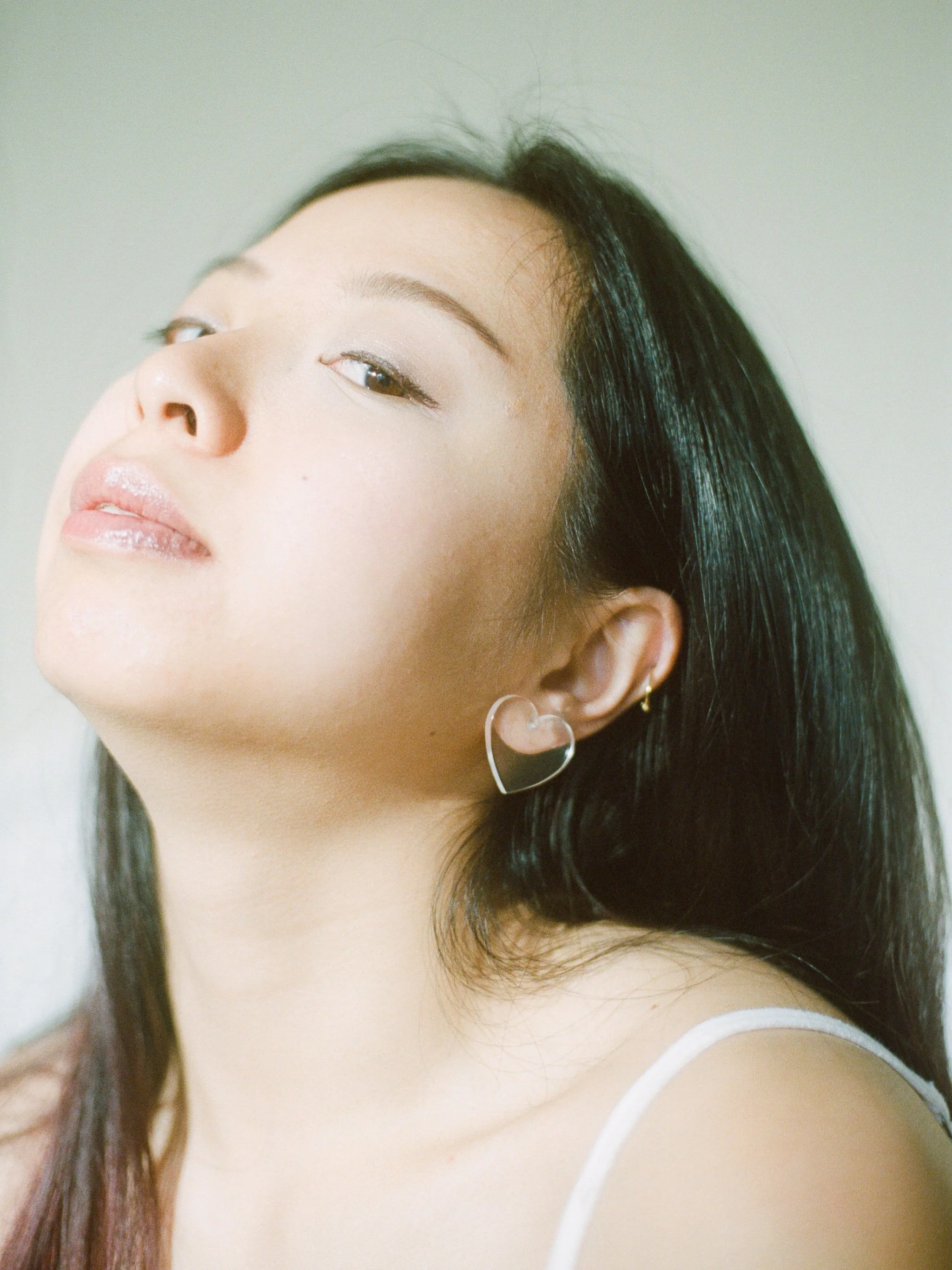 Close-up of a young woman's face with black hair, earrings, and nude makeup, looking over her shoulder against a neutral background.