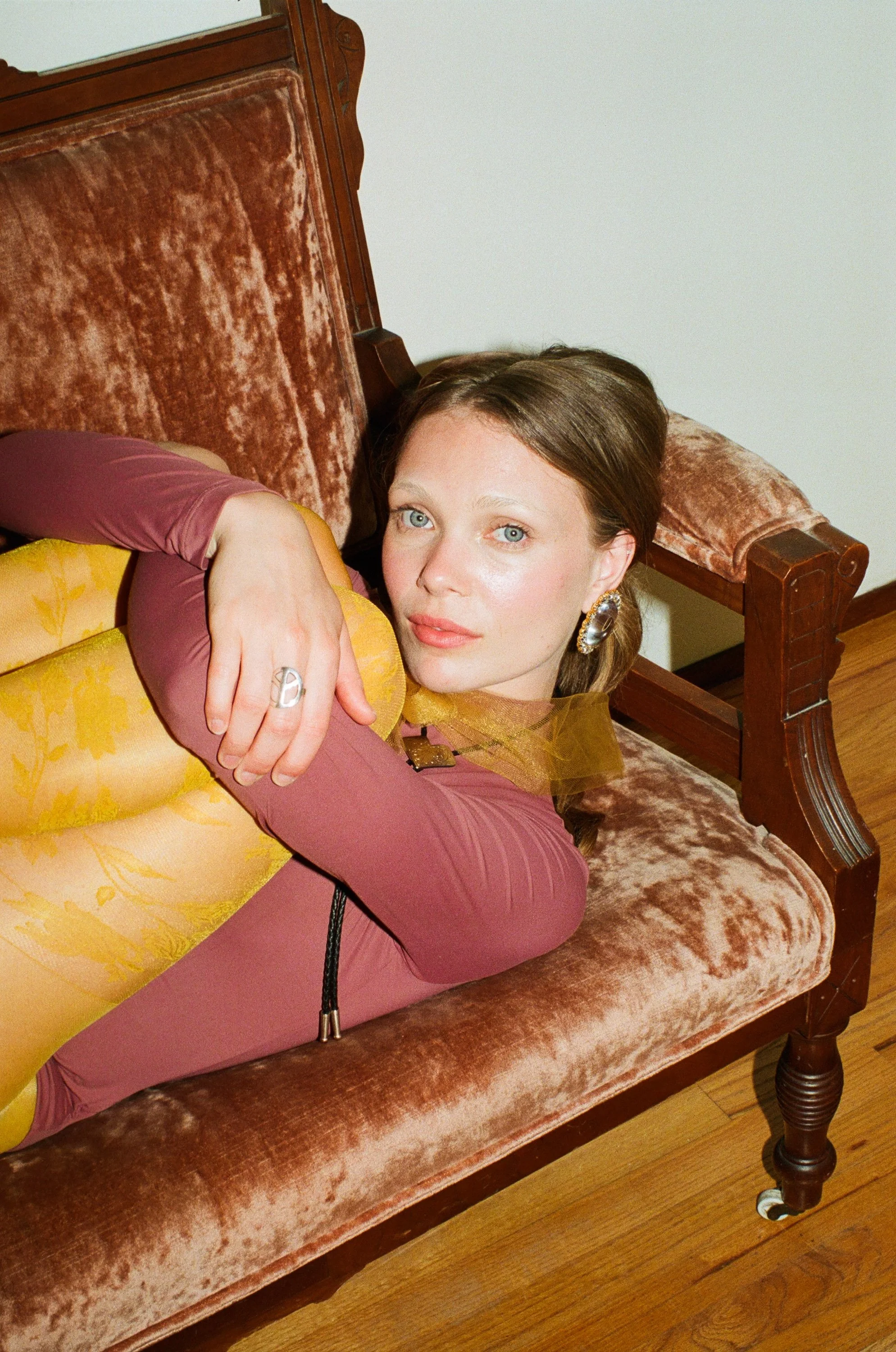 Fashion model with blue eyes wearing a pink jumpsuit with sheer sleeves, reclining on a pink velvet sofa with a floral cushion while looking at the camera.