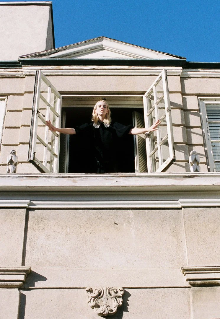 Fashion model with blonde hair standing at an open window with her arms outstretched, framed by a classic-style building against a clear blue sky.