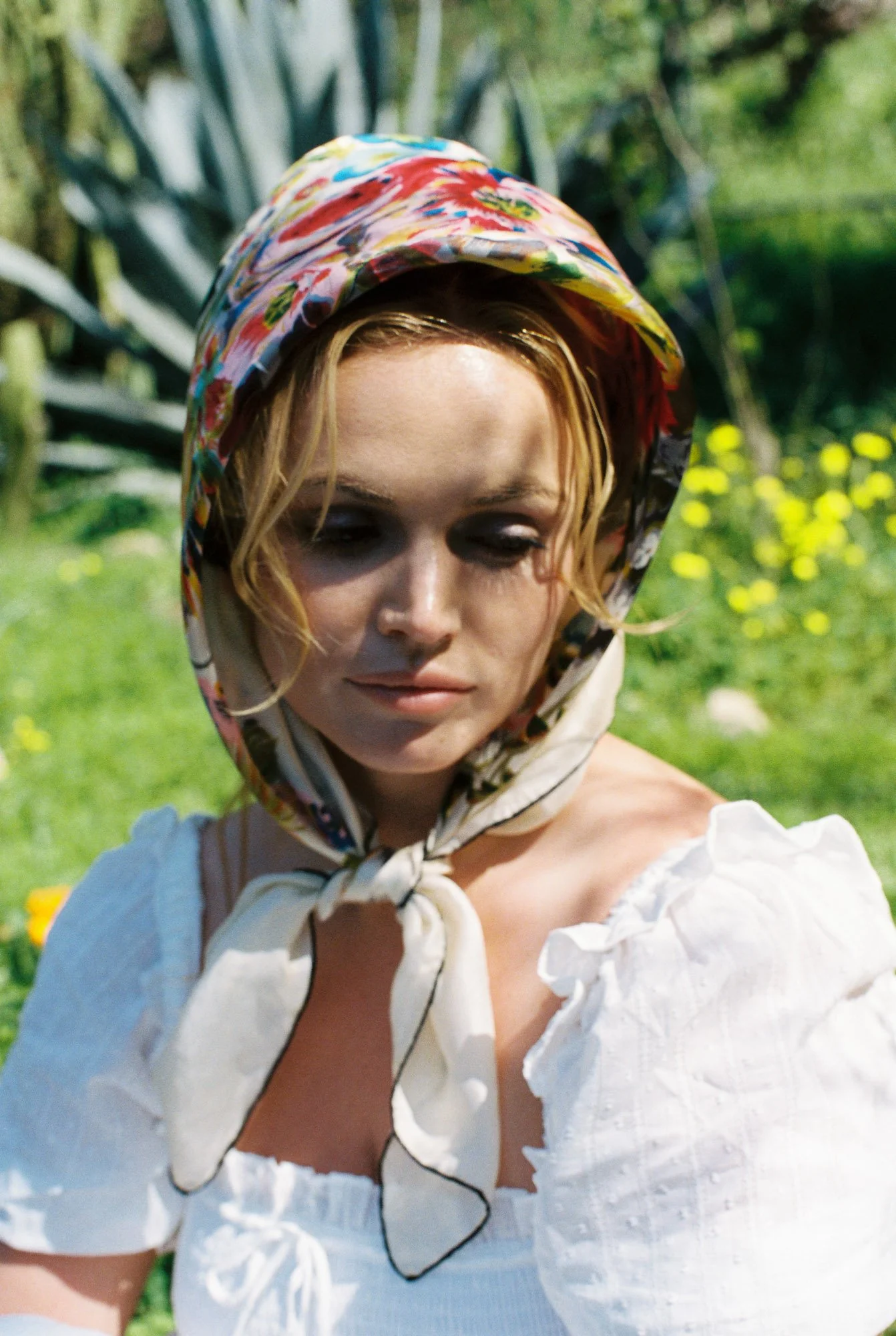 Fashion model wearing a colorful floral headscarf and a white blouse, standing outdoors in a garden with greenery and yellow flowers.