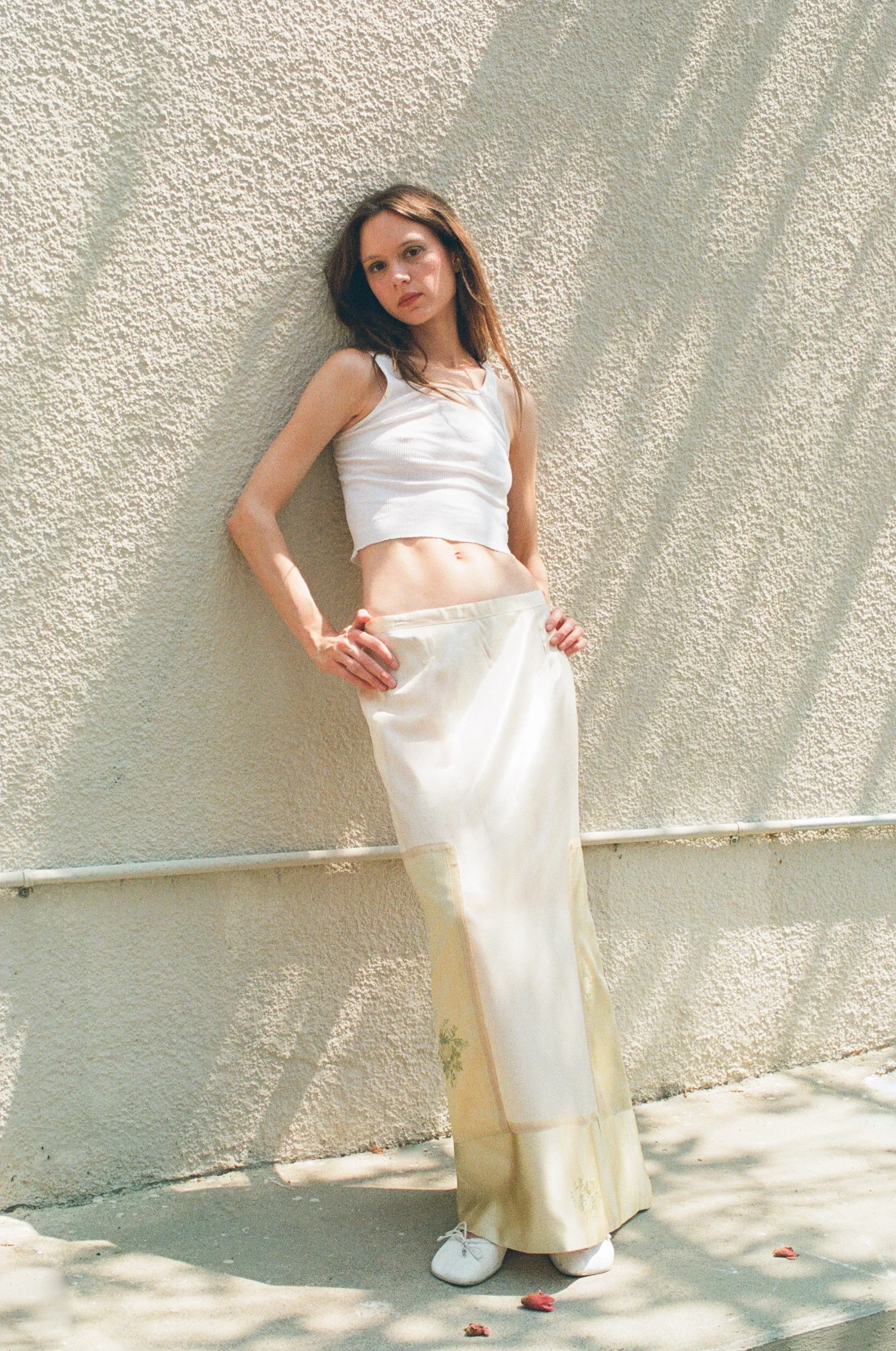 Model with brown hair wearing a white crop top and beige pants standing against a textured outdoor wall with flower petals scattered on the ground.