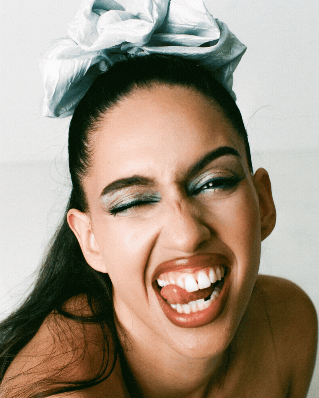 A woman with dark hair, dramatic makeup, and a large gray bow in her hair making a playful face, winking with one eye, sticking out her tongue, and showing her teeth.