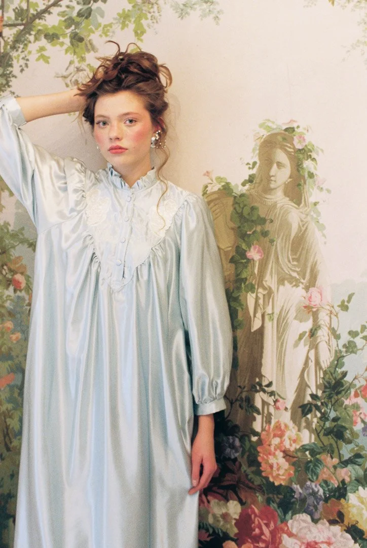Fashion model with curly hair wearing a white satin vintage dress, posing in front of a floral vintage-style mural.