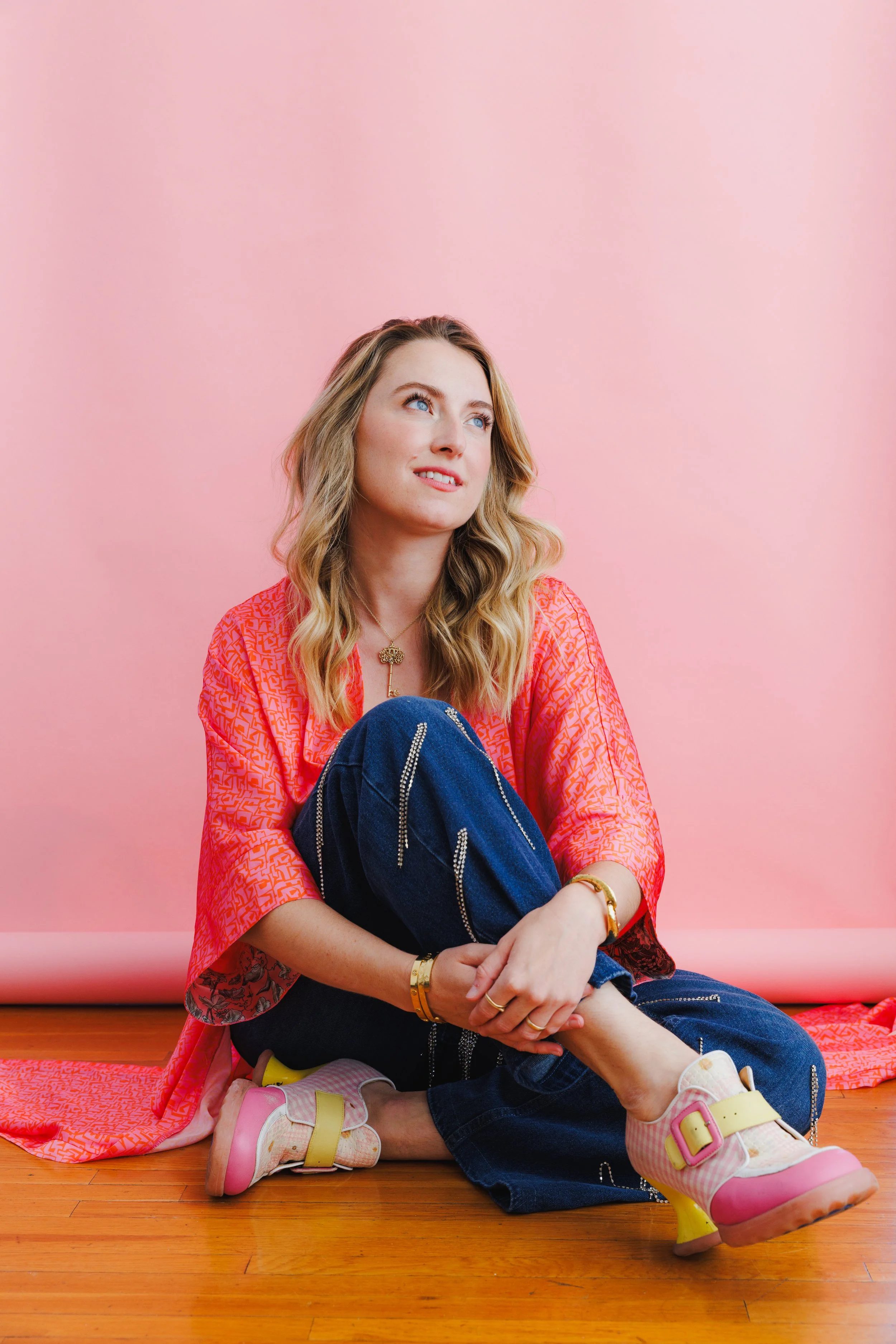 Lifestyle portrait of a woman seated casually in front of a pink backdrop, wearing a colorful red top, blue jeans, and playful platform shoes.