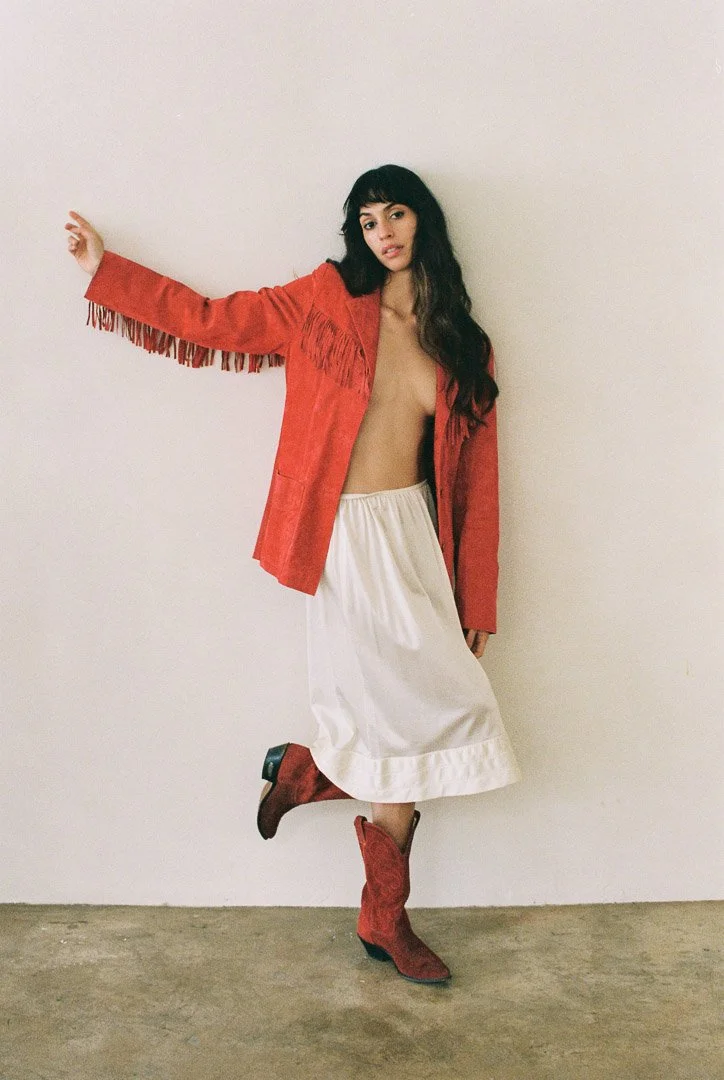 Fashion model standing against a white studio wall wearing a red jacket, cream-colored skirt, and red cowboy boots.