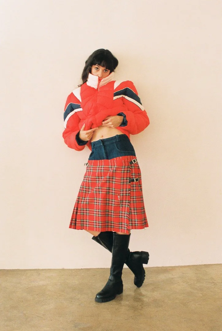 Editorial portrait of a fashion model wearing a color-block puffer jacket, red plaid skirt, and black knee-high boots against a white studio wall.
