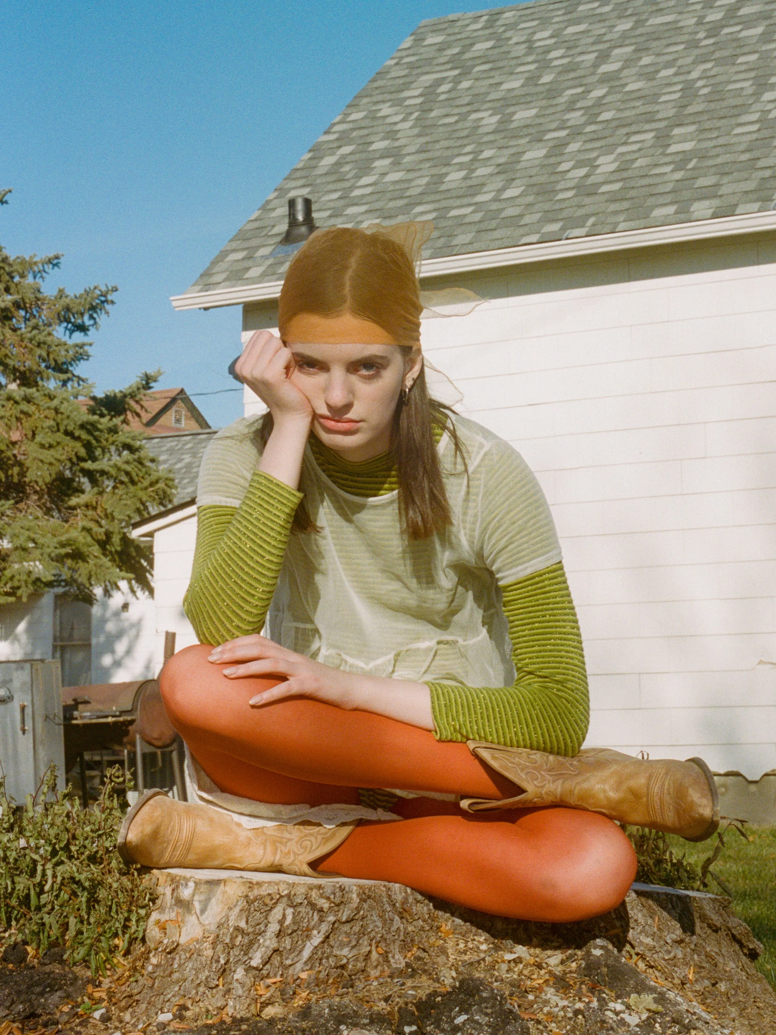 Fashion model with red hair wearing a striped green and yellow top, tan cowboy boots, and a sheer yellow hair scarf, sitting on a tree stump outdoors with a white house in the background.