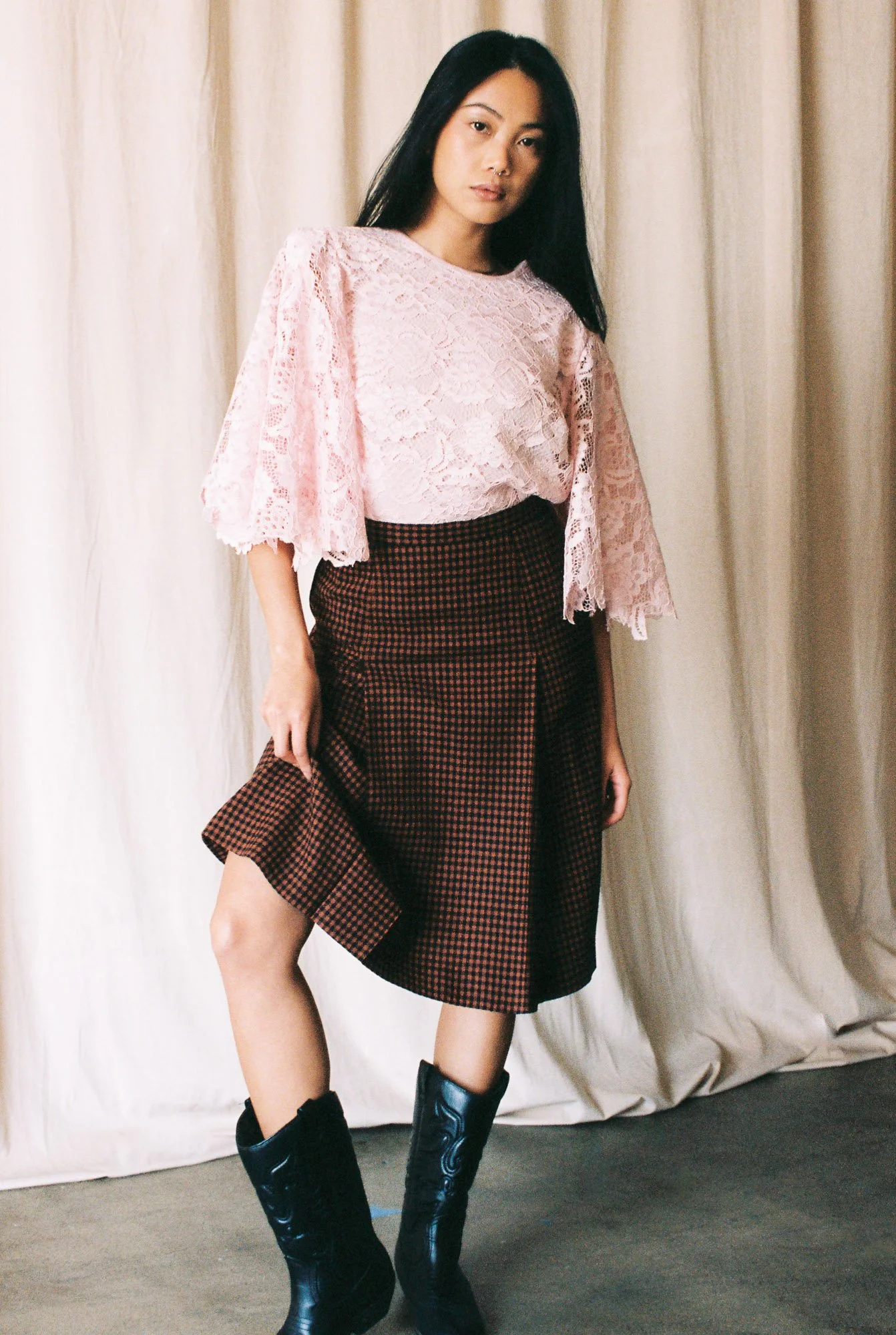 A woman with long black hair wearing a pink lace top, a checkered brown and red skirt, and black cowboy boots standing in front of a cream-colored curtain.