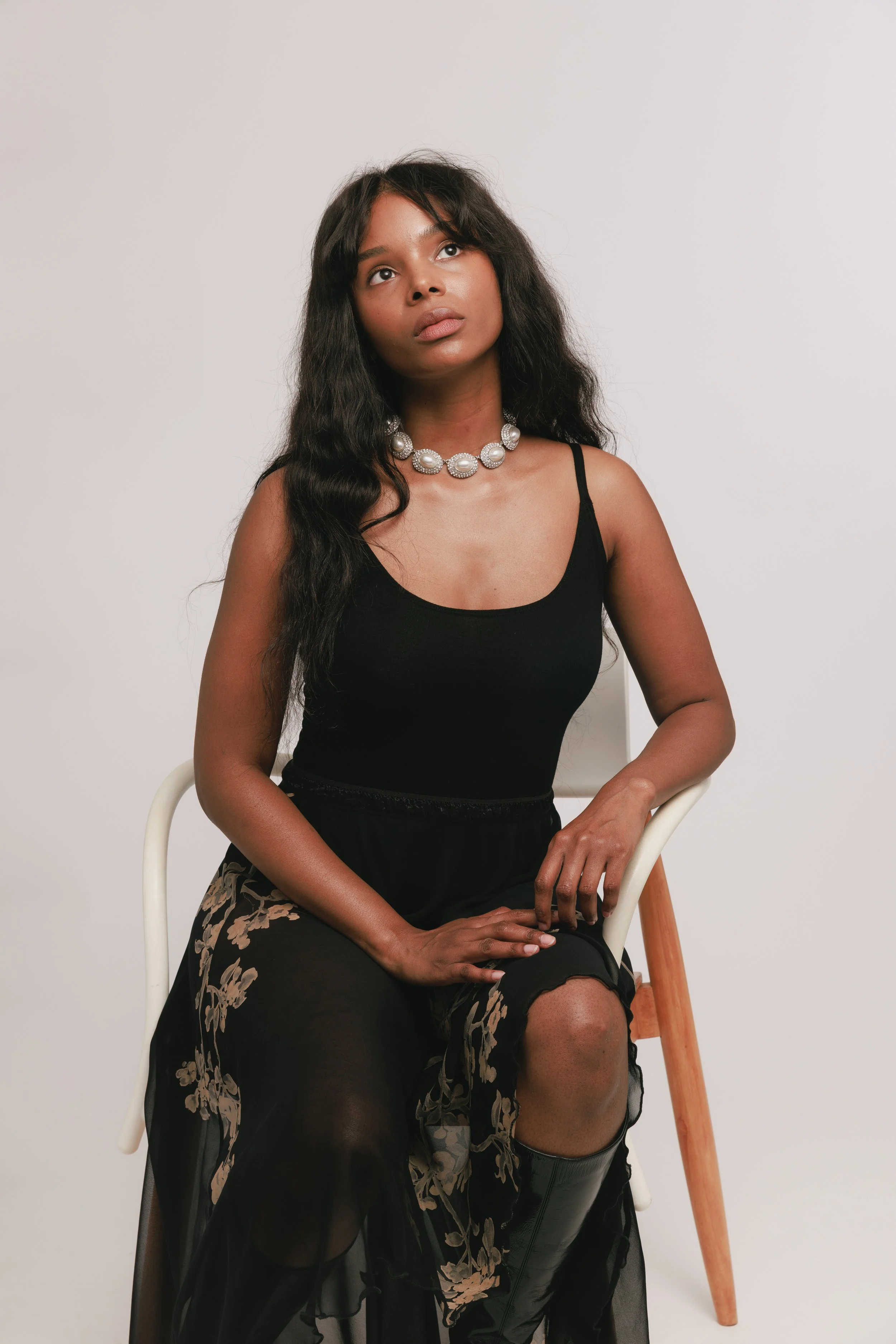 Fashion model with long wavy black hair wearing a black tank top, a sheer black skirt with floral embroidery, a pearl necklace, and black boots, seated on a white chair against a white background while looking upward.